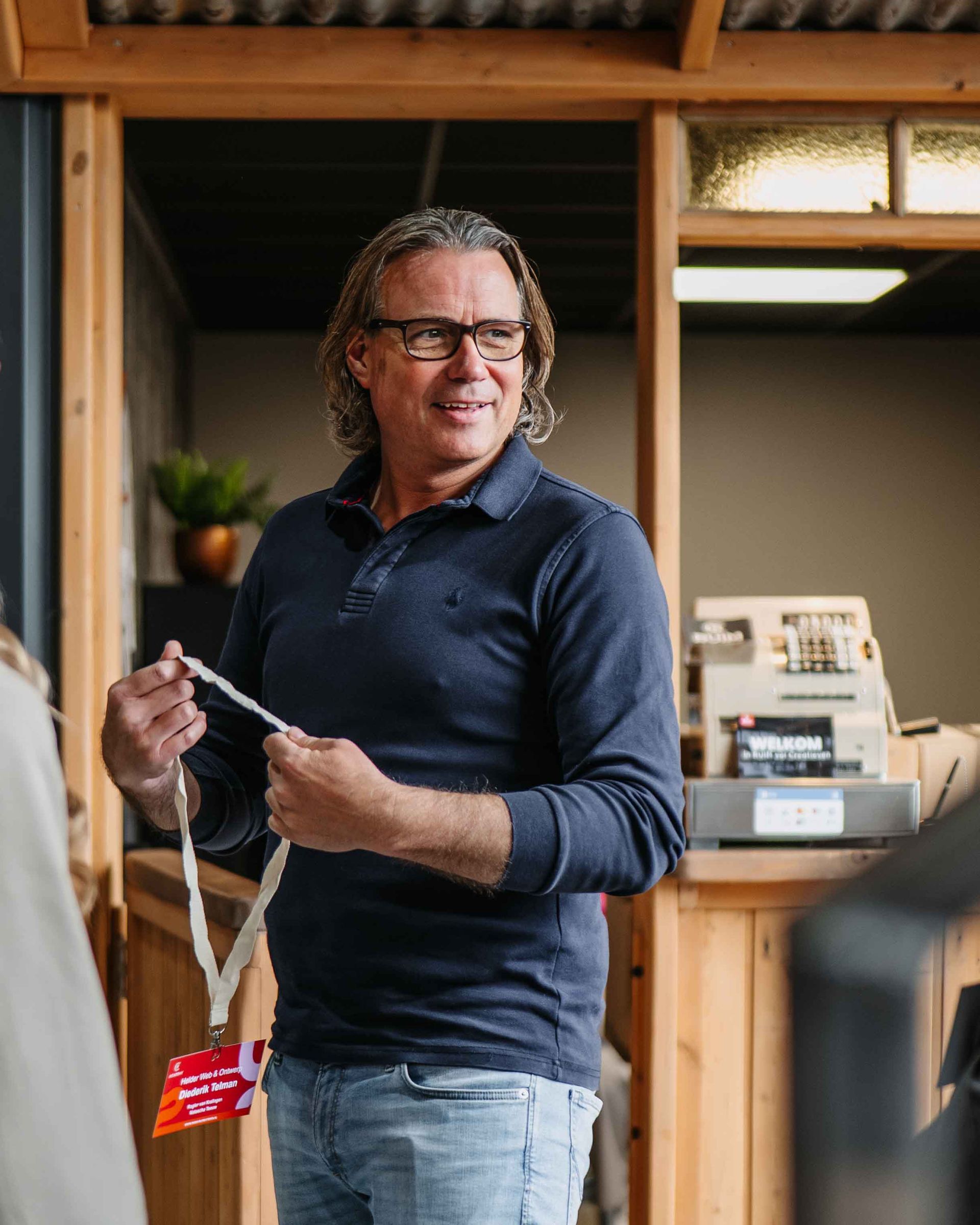 Een persoon met lang haar en een bril houdt een sleutelkoord vast terwijl hij in een kamer met houten lambrisering staat.
