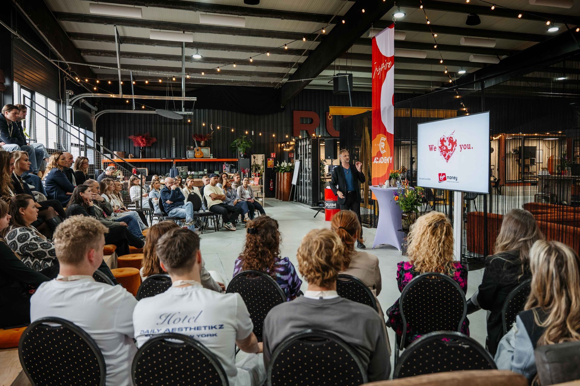 Een persoon geeft een presentatie aan een publiek dat zit in een ruime, industriële ruimte waar een logo op een groot scherm wordt weergegeven.