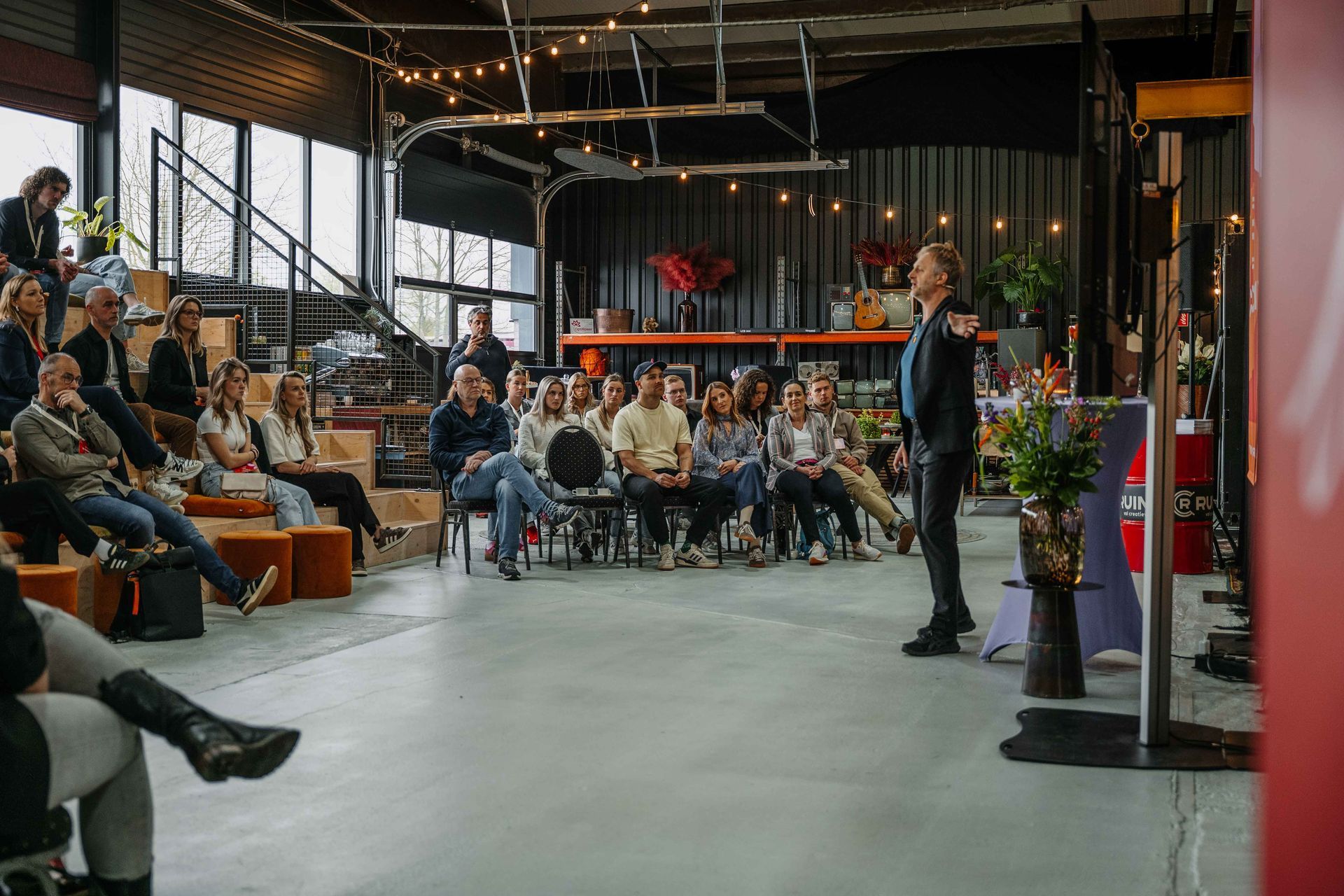 Een spreker spreekt een zittend publiek toe in een lichte, industriële ruimte met houten tribune.
