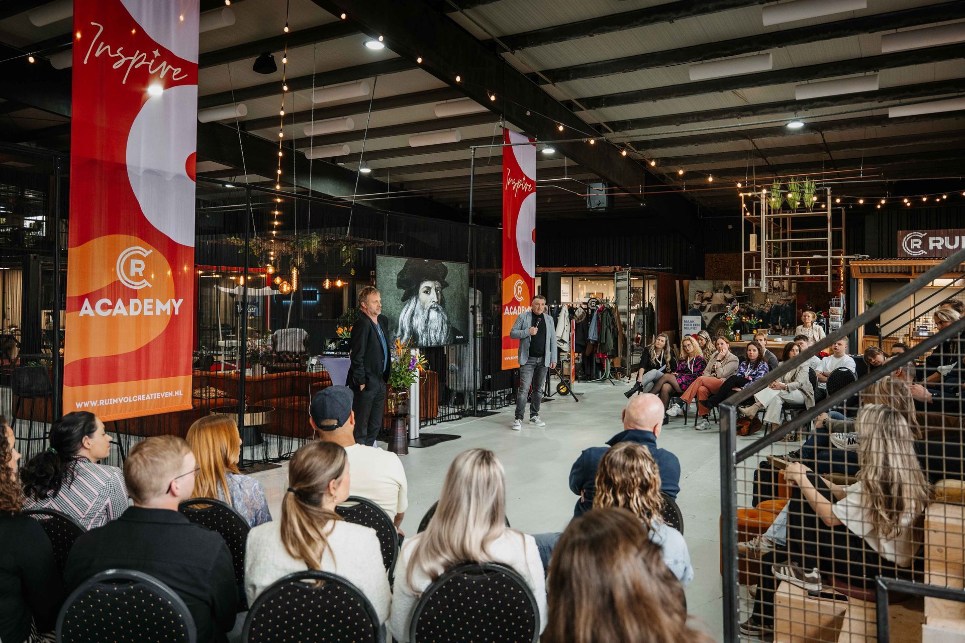 Een spreker geeft een presentatie voor een publiek in een grote, industriële zaal die is versierd met oranje banners van de Academie.