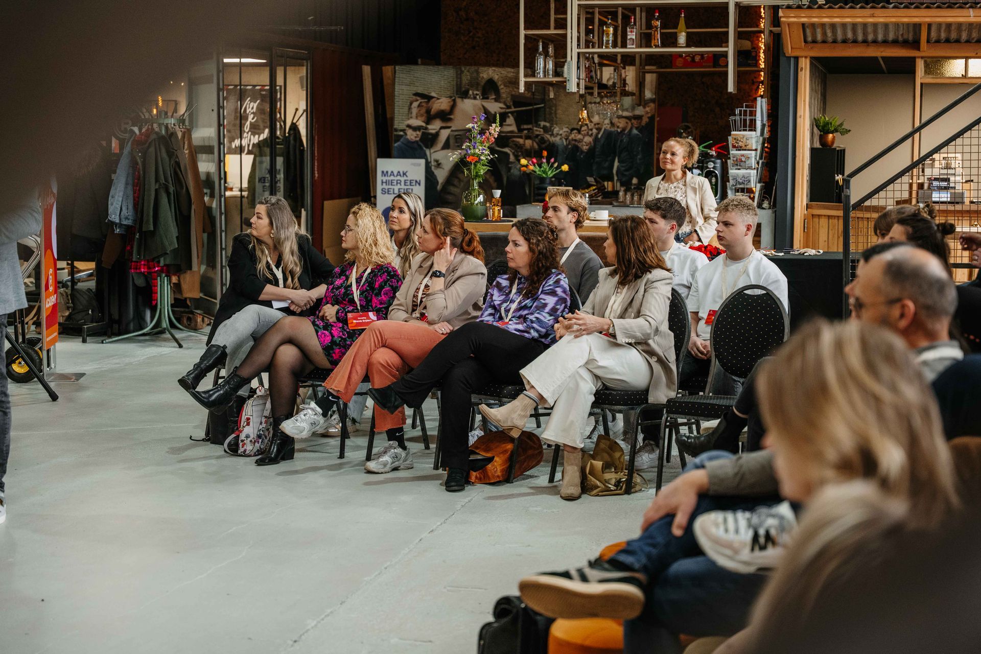 Een groep mensen zit in een studio in industriële stijl, met hun gezicht naar voren gericht, en luistert naar een seminar of presentatie.
