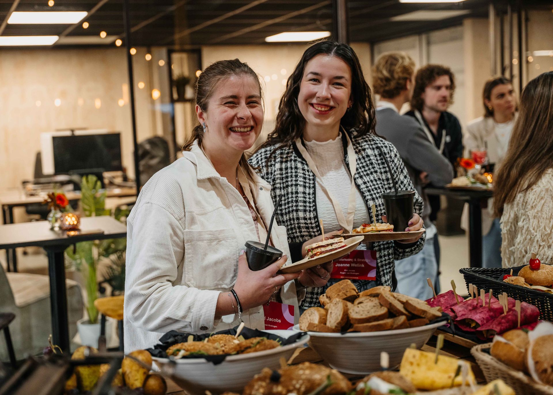 Twee mensen glimlachen en houden snacks vast tijdens een informeel sociaal evenement binnenshuis, met eetkraampjes op de voorgrond.