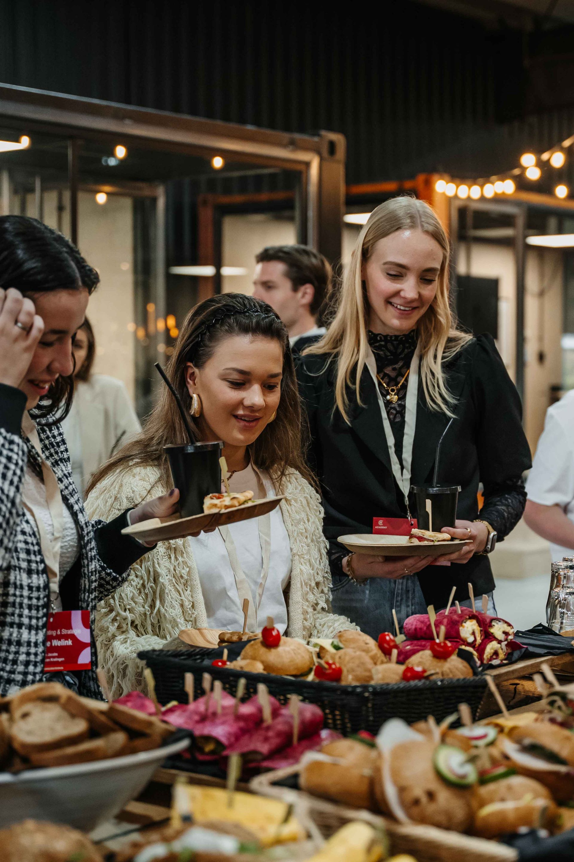 Drie mensen lachend terwijl ze kleine hamburgers en snacks uitkiezen van een buffet op een evenement binnenshuis.