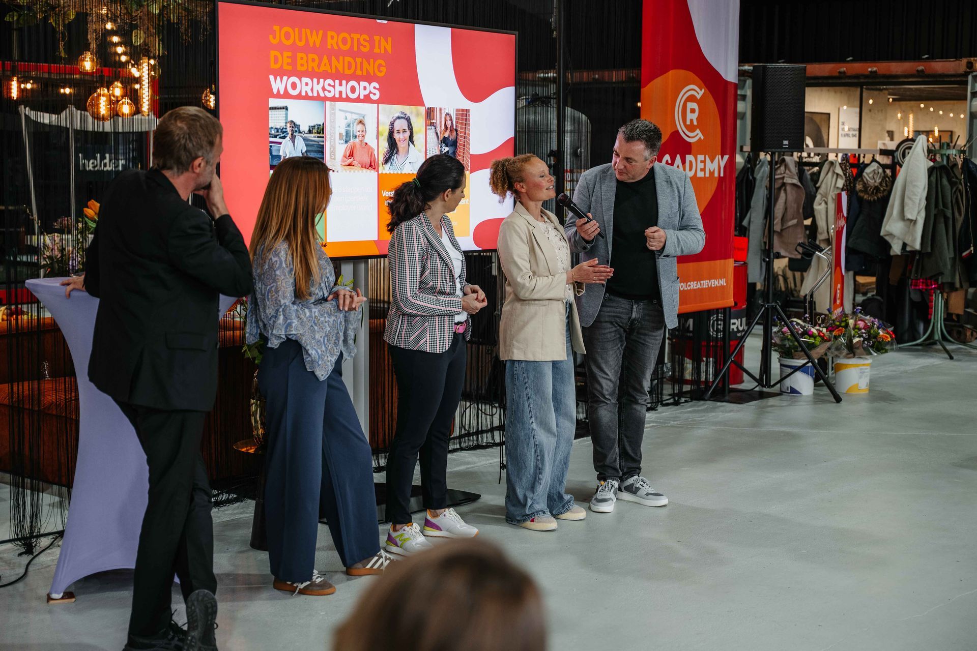 Een panel van vijf personen staat op een podium voor een rood scherm, waarbij één persoon in een microfoon spreekt.