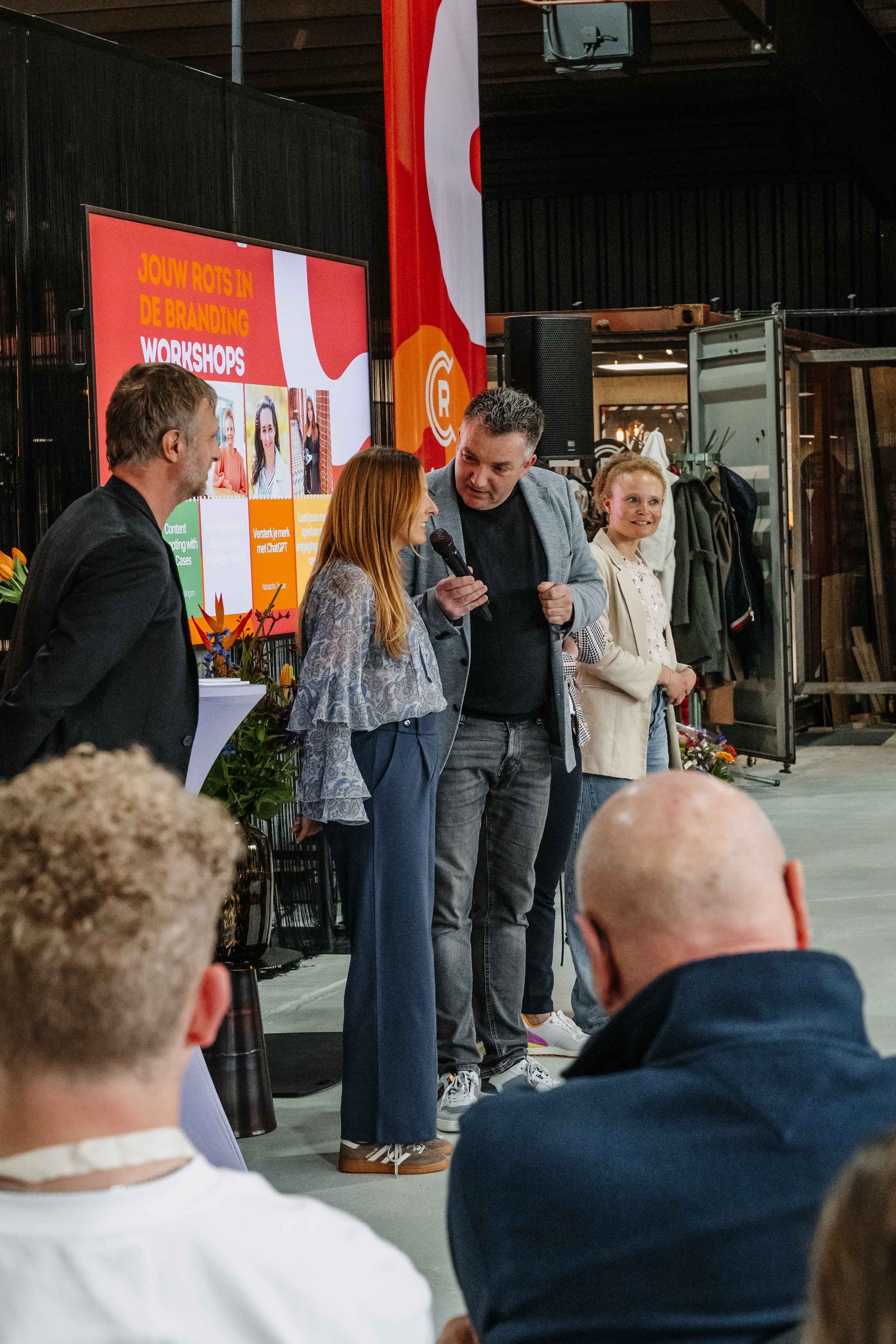 Een groep mensen staat op een podium tijdens een evenement binnenshuis, voor een kleurrijk spandoek met tekst.
