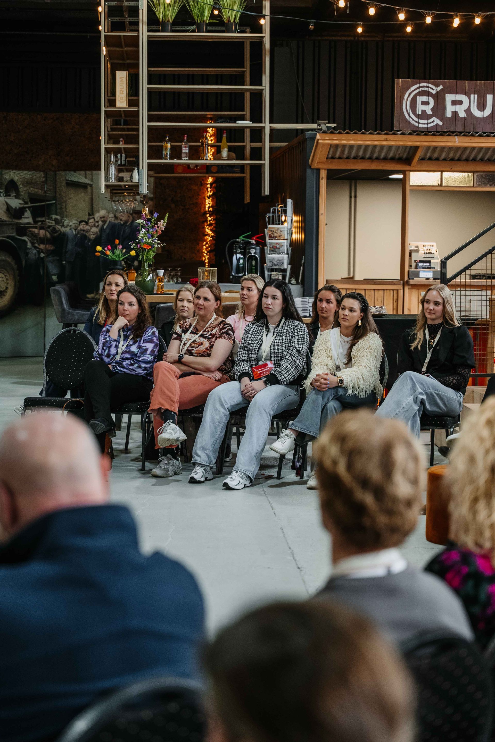 Een groep mensen zit op stoelen in een evenementenruimte in magazijnstijl en luistert naar een presentatie.