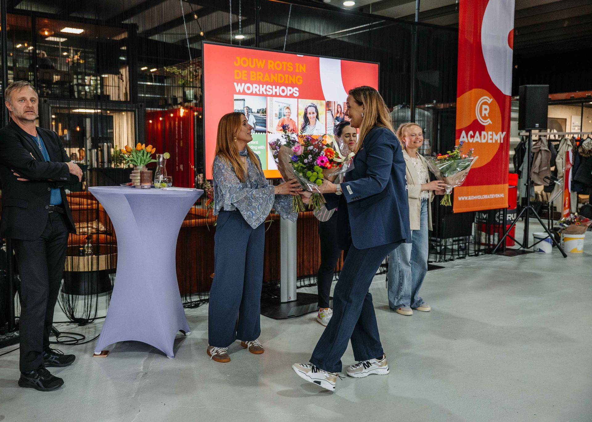 Tijdens een evenement staan ​​mensen op een podium, waarbij één persoon boeketten bloemen uitdeelt aan anderen voor een achtergrond.