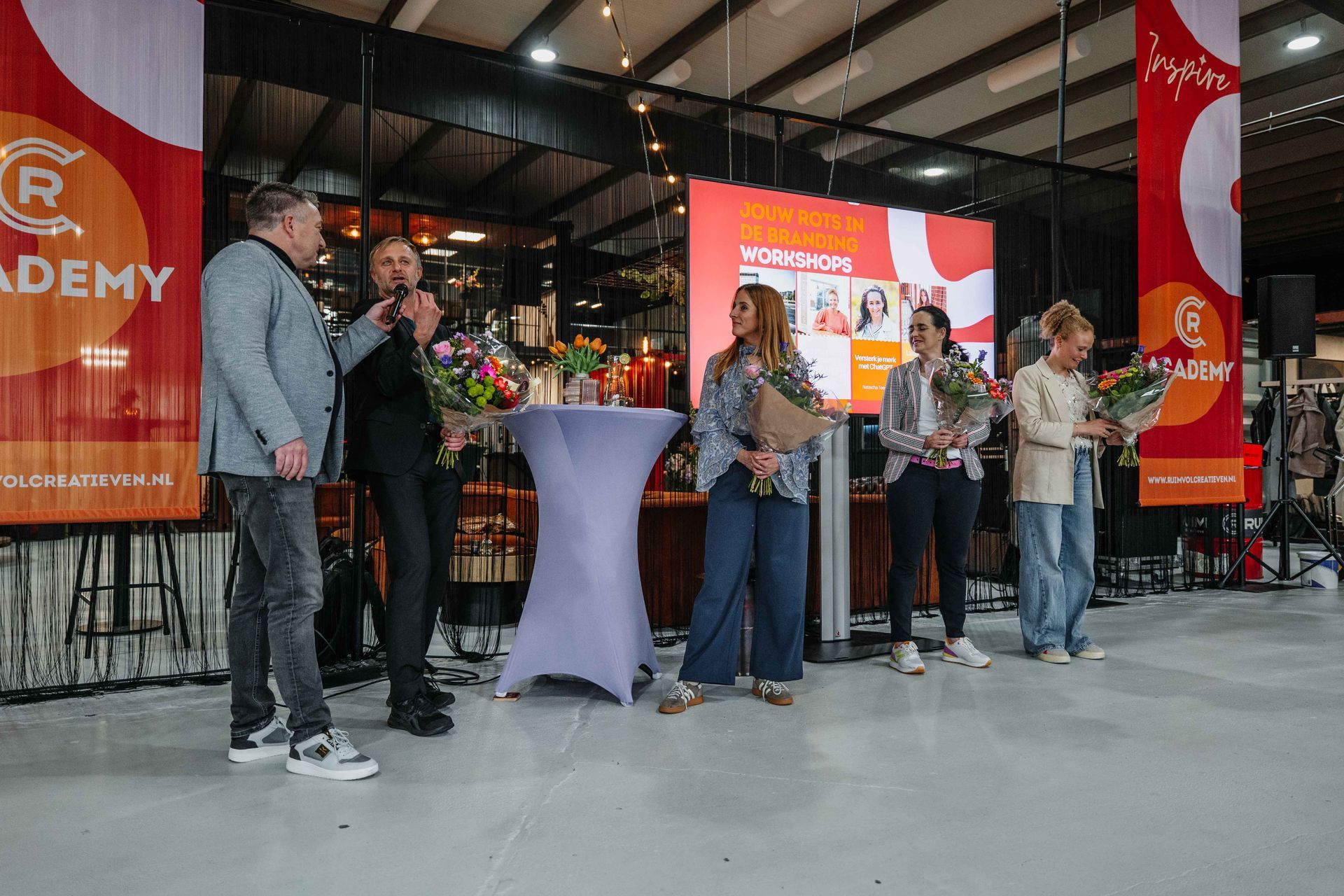 Een groep mensen staat op een podium tijdens een evenement binnenshuis, waarvan drie boeketten vasthouden voor een oranje scherm met het bedrijfslogo.
