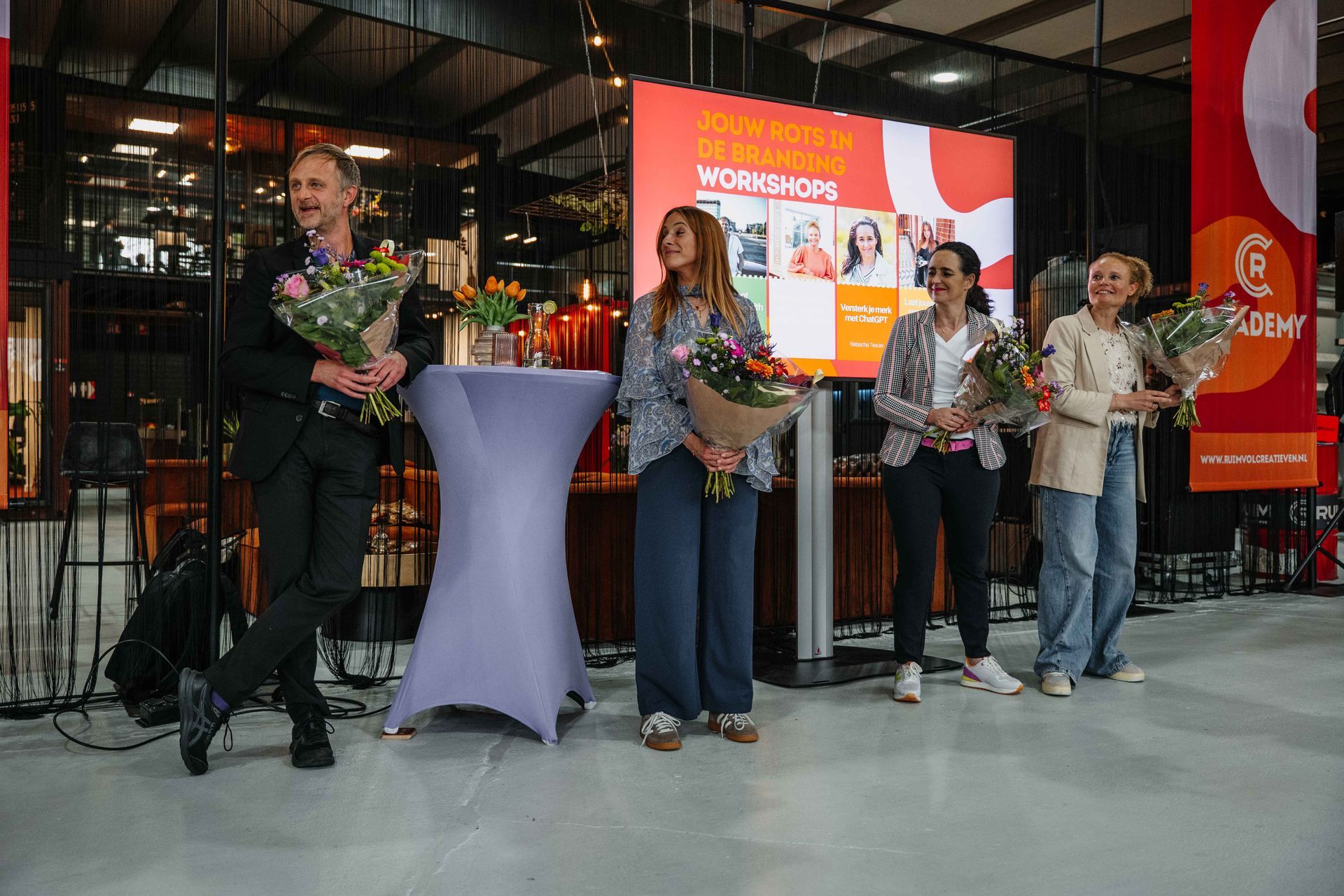 Vier mensen staan ​​op een podium met boeketten bloemen in hun handen tijdens een evenement met een groot digitaal scherm op de achtergrond.