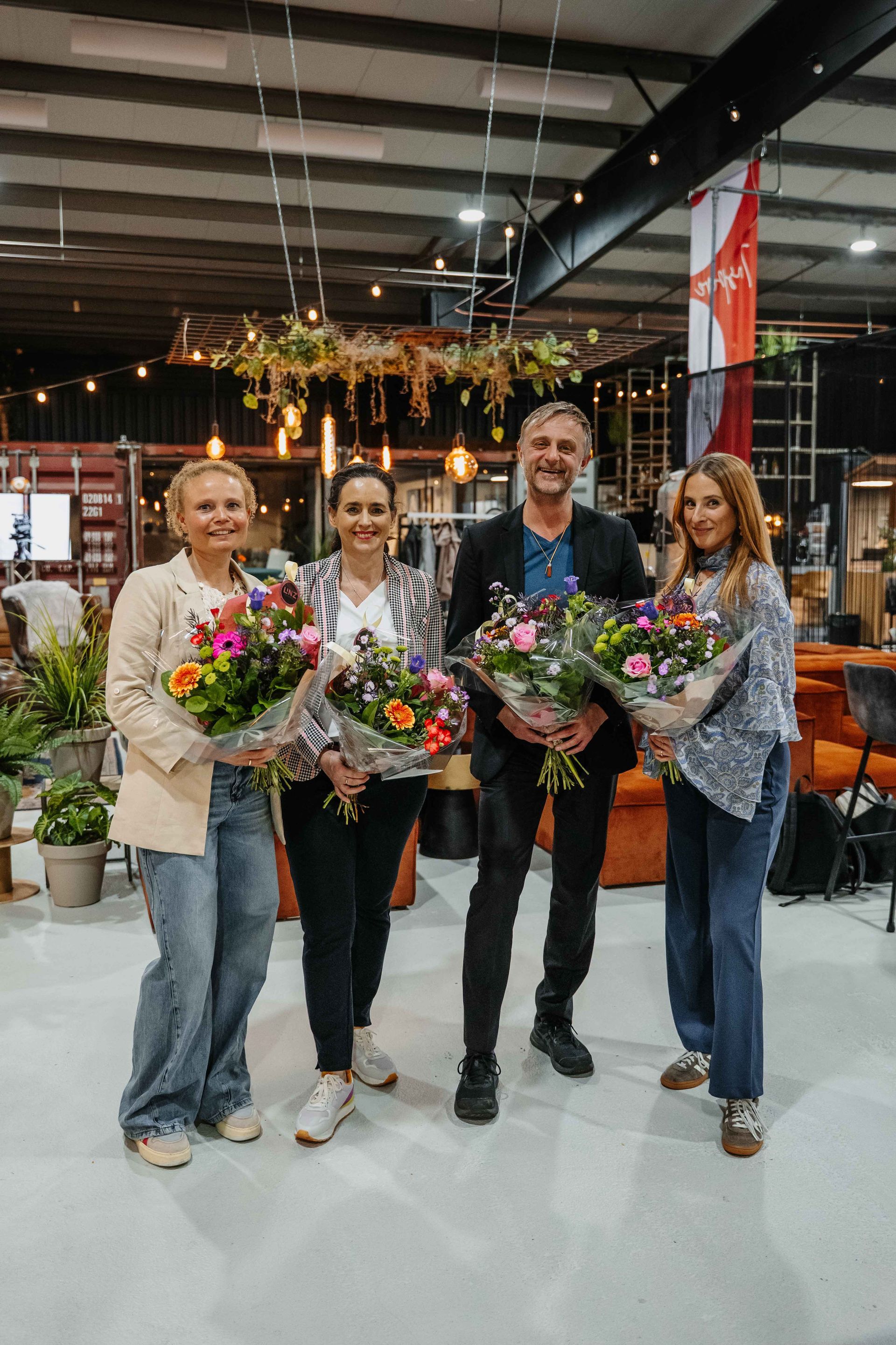 Vier mensen staan ​​samen in een werkplaats, lachend en met boeketten kleurrijke bloemen in hun handen.