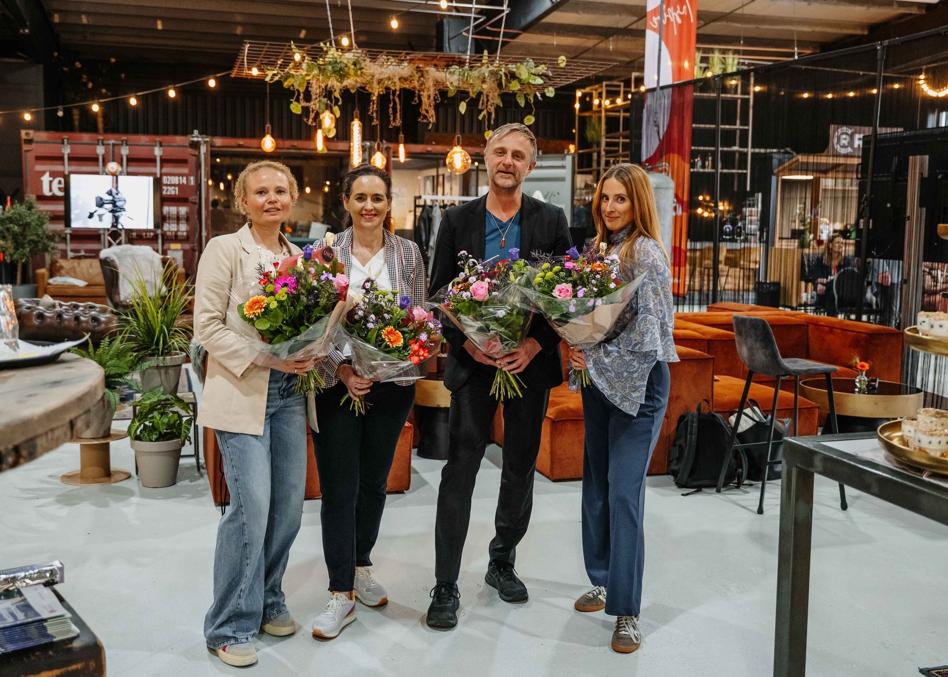 Vier mensen staan met boeketten bloemen in hun handen in een showroom in industriële stijl, compleet met meubels en verlichting.