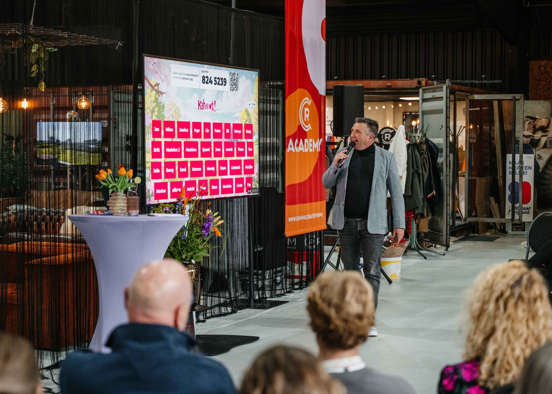 Een spreker geeft een presentatie in een zaal die eruitziet als een pakhuis, staand naast een spandoek met een raster van rode vierkanten en een publiek.