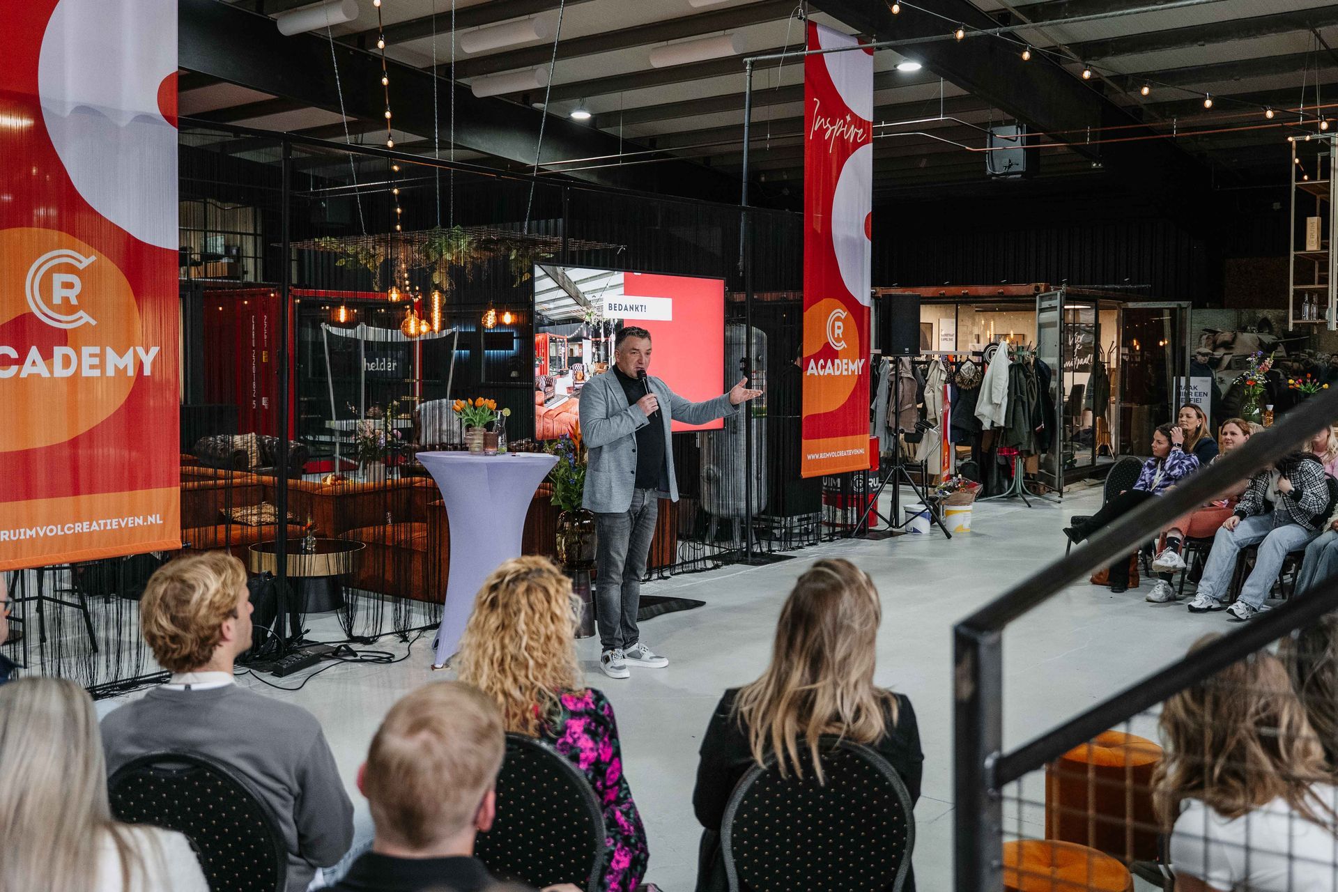 Een spreker staat op een podium en spreekt een publiek toe in een grote, industriële zaal die versierd is met rode Academy-banners.
