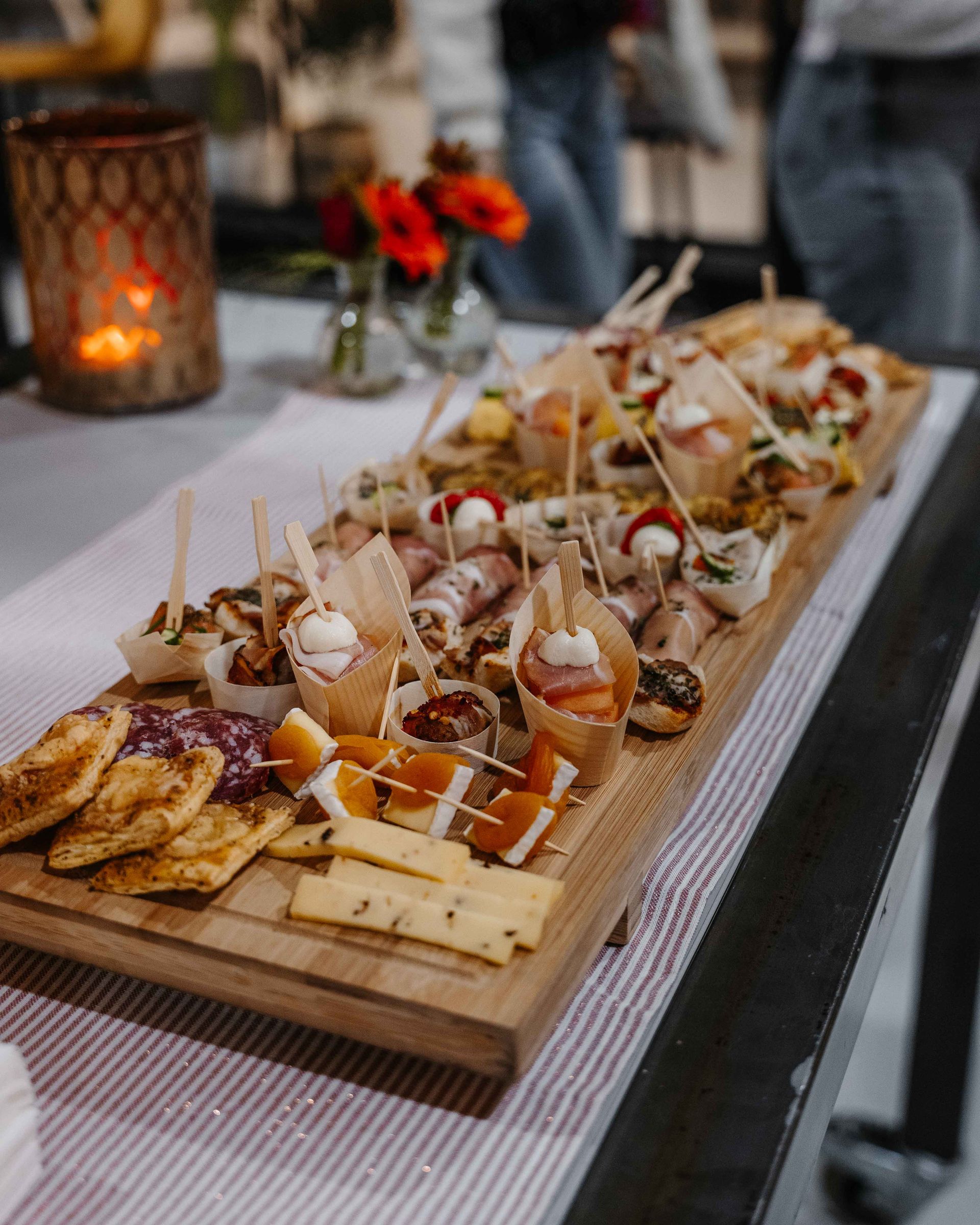 Een lange houten plank vol met diverse hapjes, waaronder kaas, crackers en verpakte vleessnacks, op een tafel.