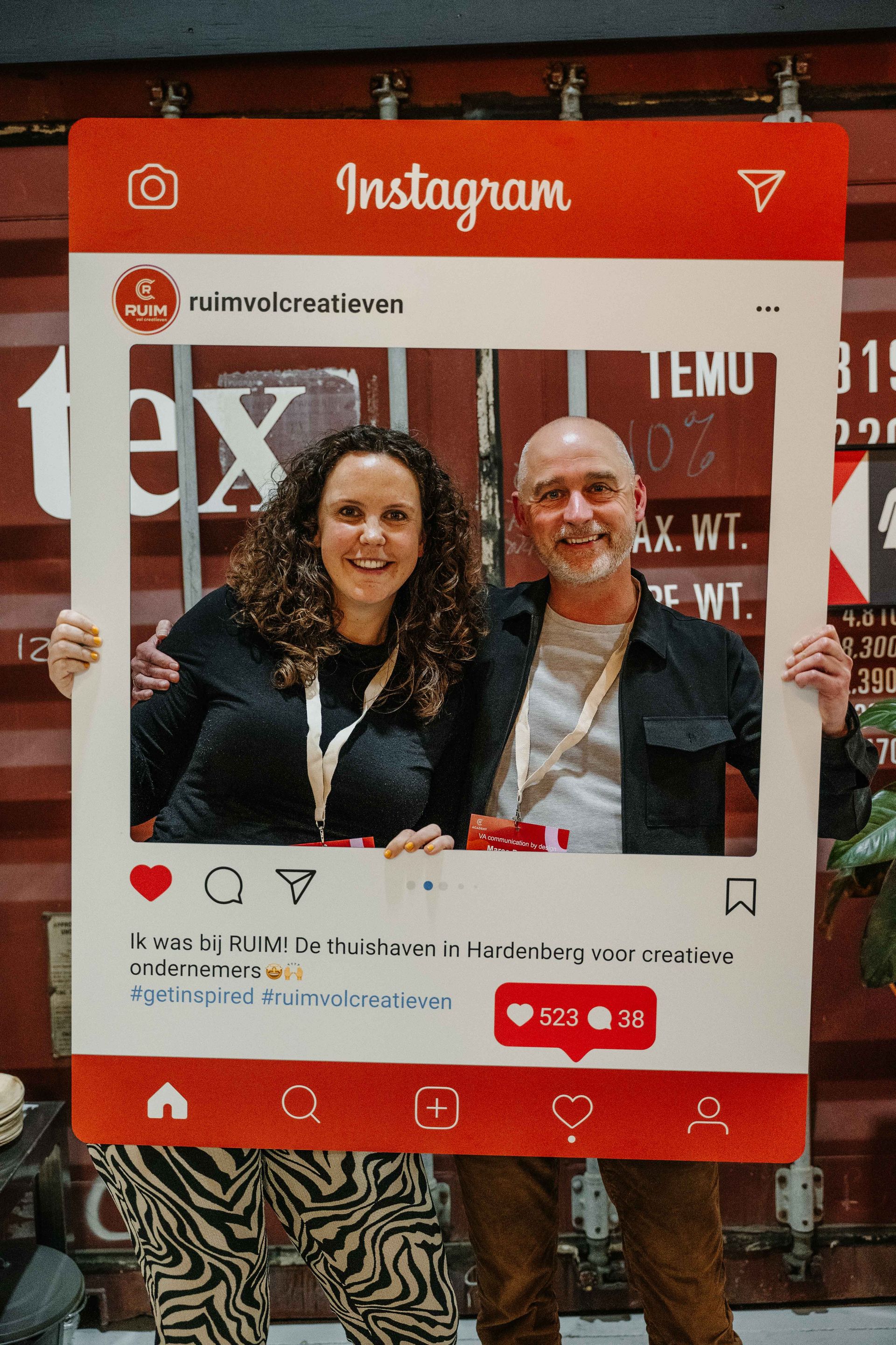 Twee mensen glimlachen in een grote, rode kartonnen fotolijst met Instagram-thema, tegen een industriële containerwand.