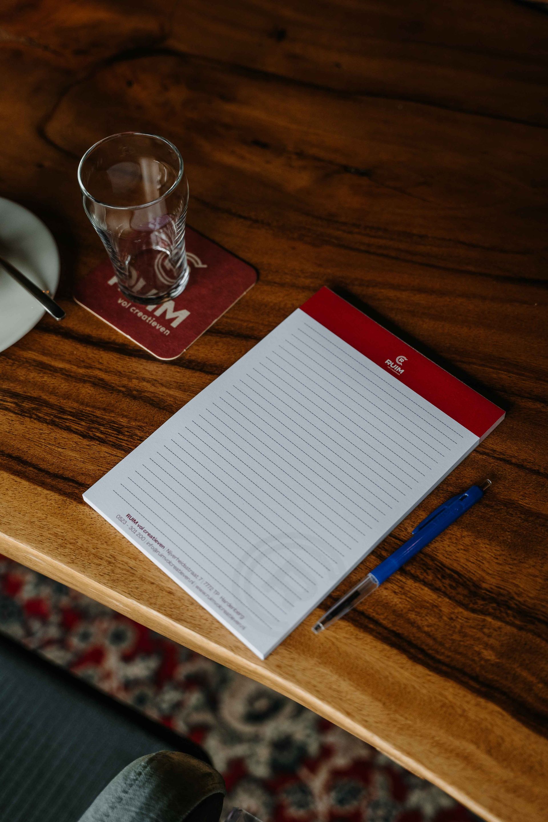 Een notitieblok met een rode koptekst, een blauwe pen en een glas water op een houten tafel.