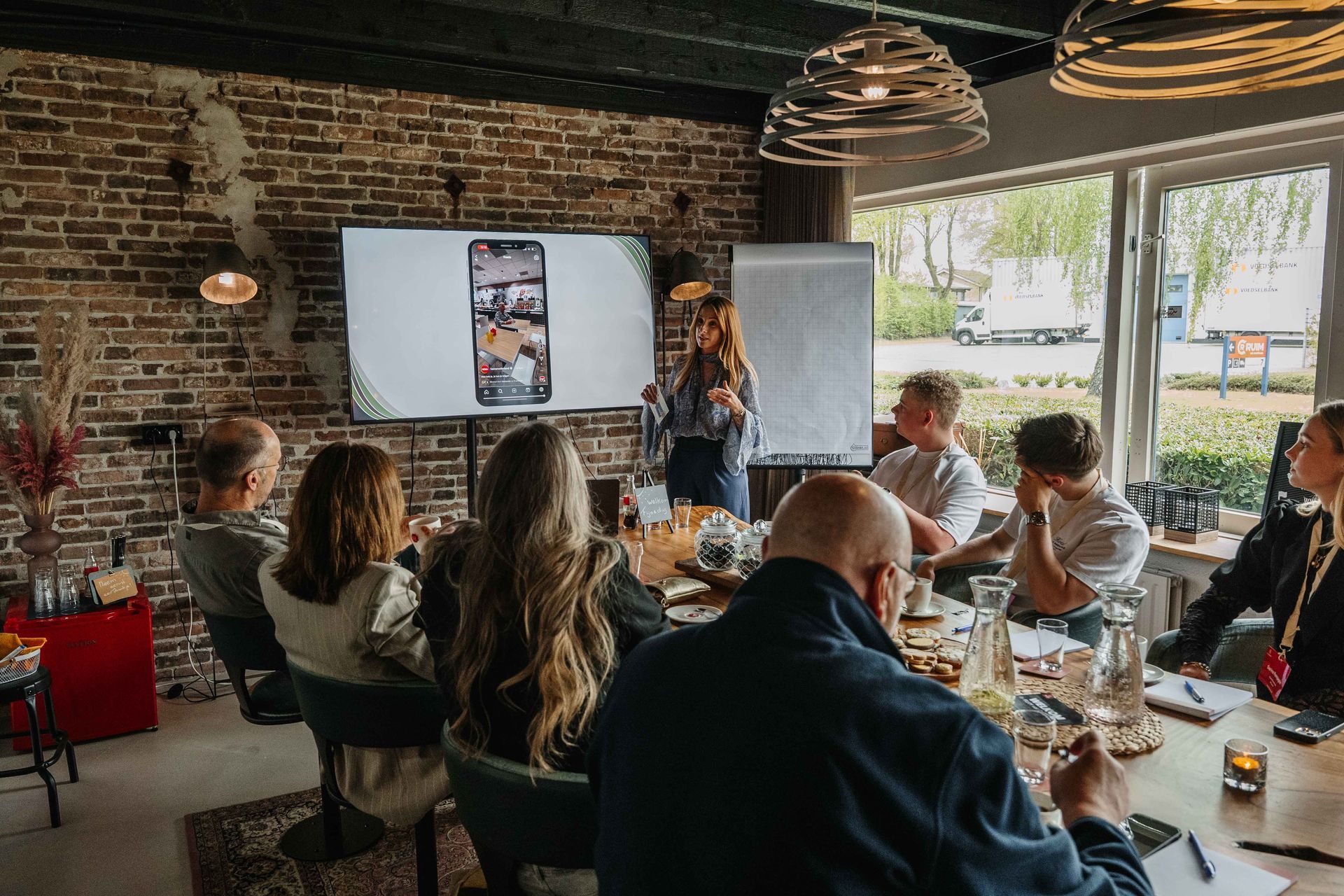 Een groep mensen zit rond een lange houten tafel in een kamer met bakstenen muren en kijkt naar een presentatie op een groot scherm.