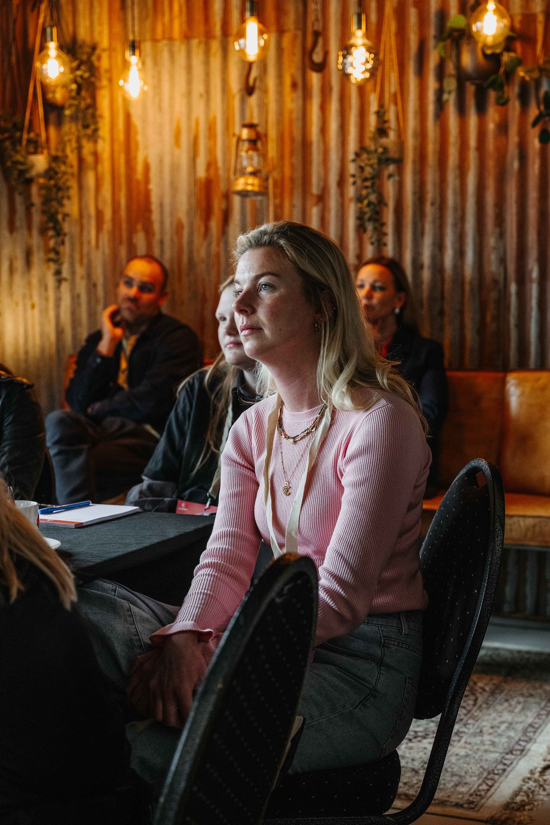 Mensen zitten in een schemerig verlichte, rustieke ruimte met golfplaten wanden en hanglampen en luisteren naar een presentatie.