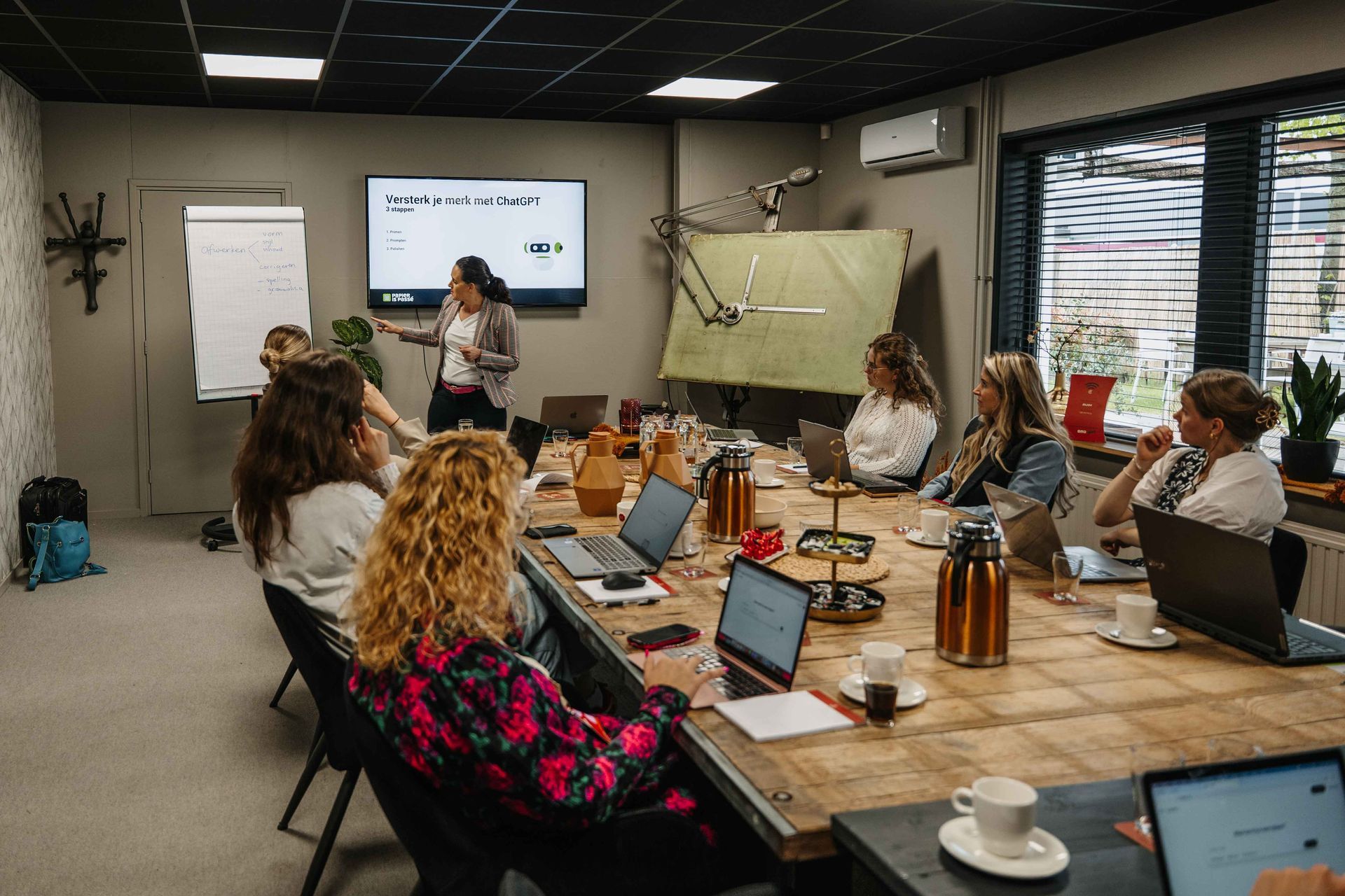 Een spreker staat voor een scherm in een vergaderruimte, terwijl een groep mensen rond een houten tafel zit te luisteren.