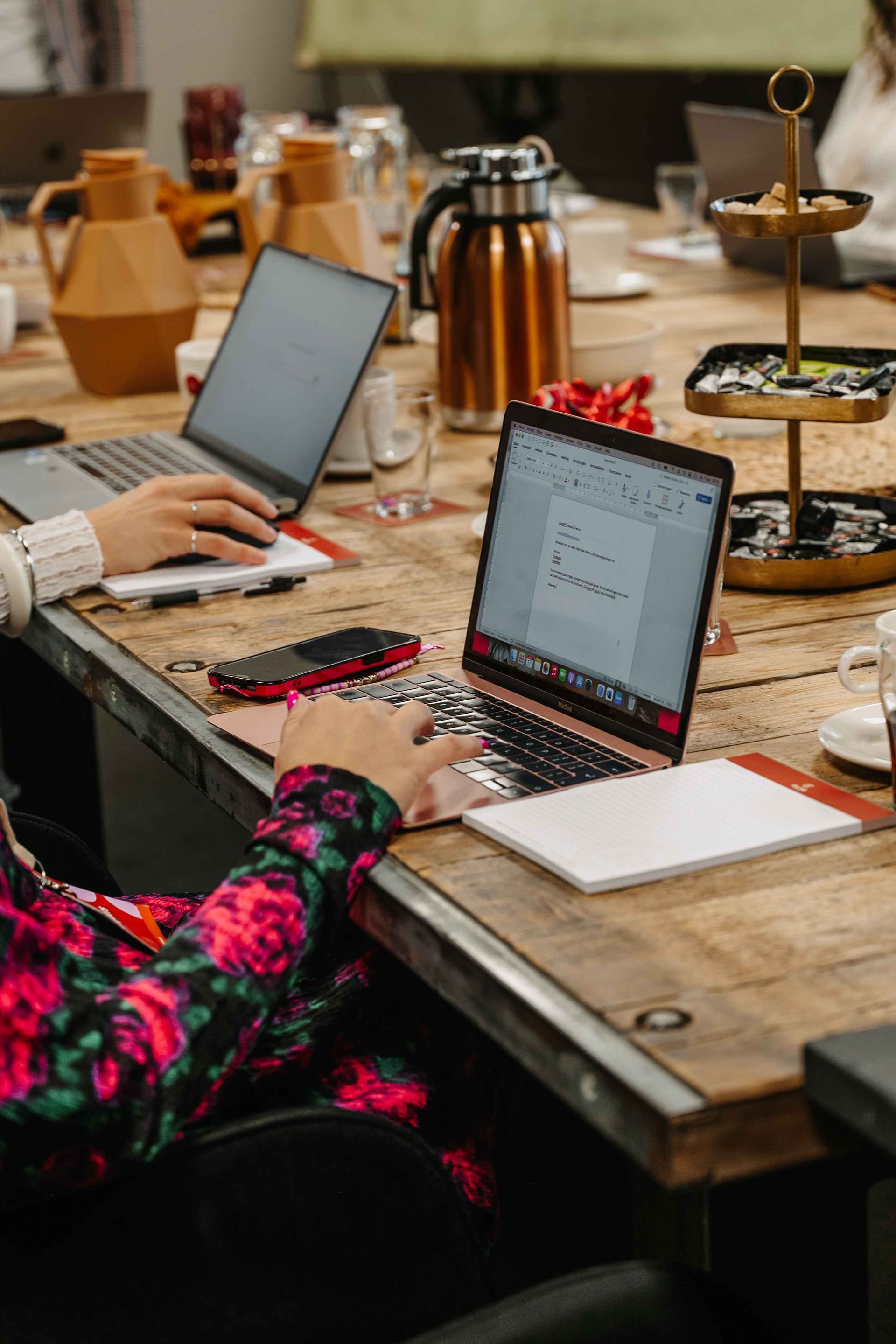 Een persoon in een bloemenhemd typt op een laptop aan een rustieke houten tafel tijdens een vergadering met collega's en drankjes.