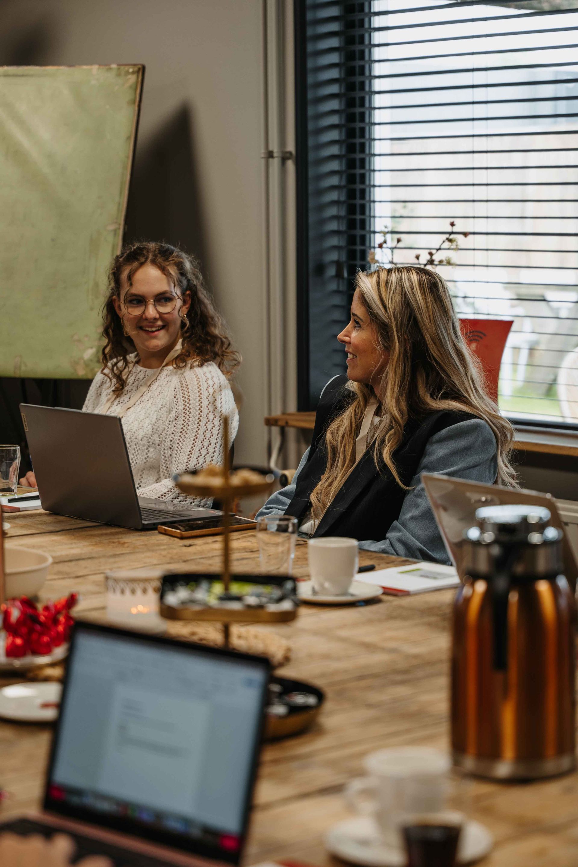 Twee mensen zitten aan een houten tafel in een kantooromgeving en werken op laptops, met drankjes in de buurt.