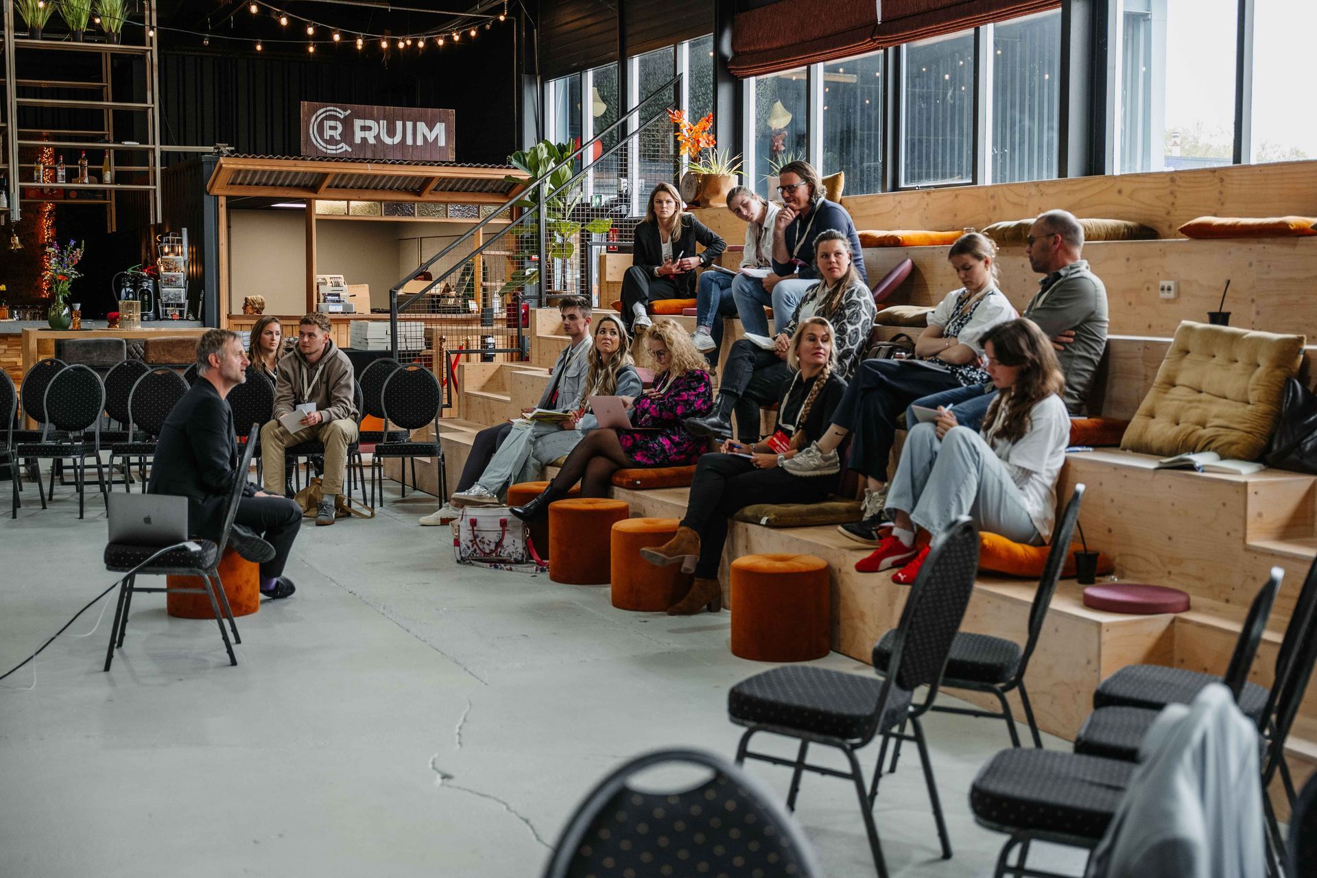 Een spreker spreekt een groep mensen toe die op houten trappen en stoelen zitten in een moderne, goed verlichte binnenruimte.