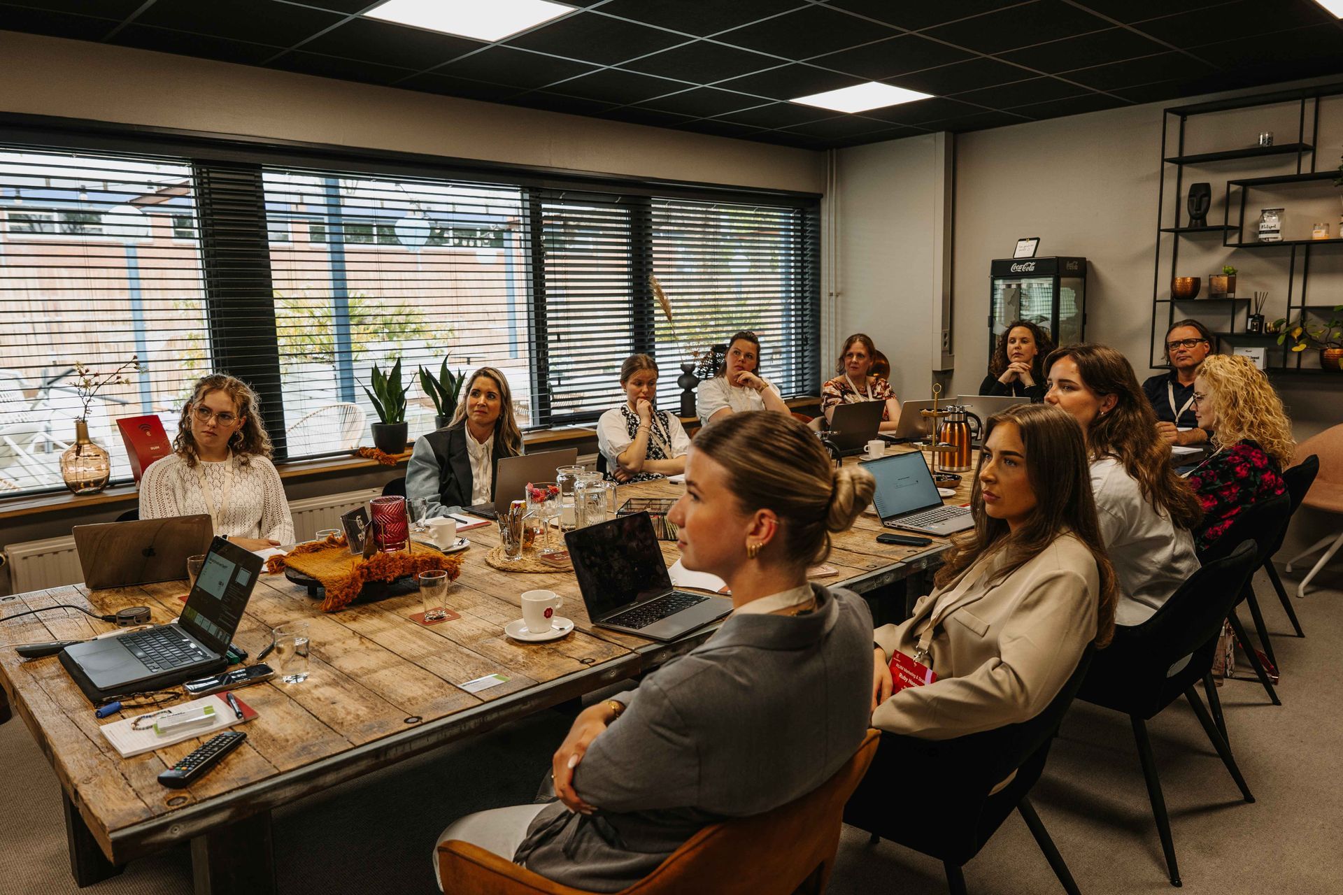 Een groep mensen zit rond een grote houten vergadertafel in een modern kantoor en luistert naar een presentatie.