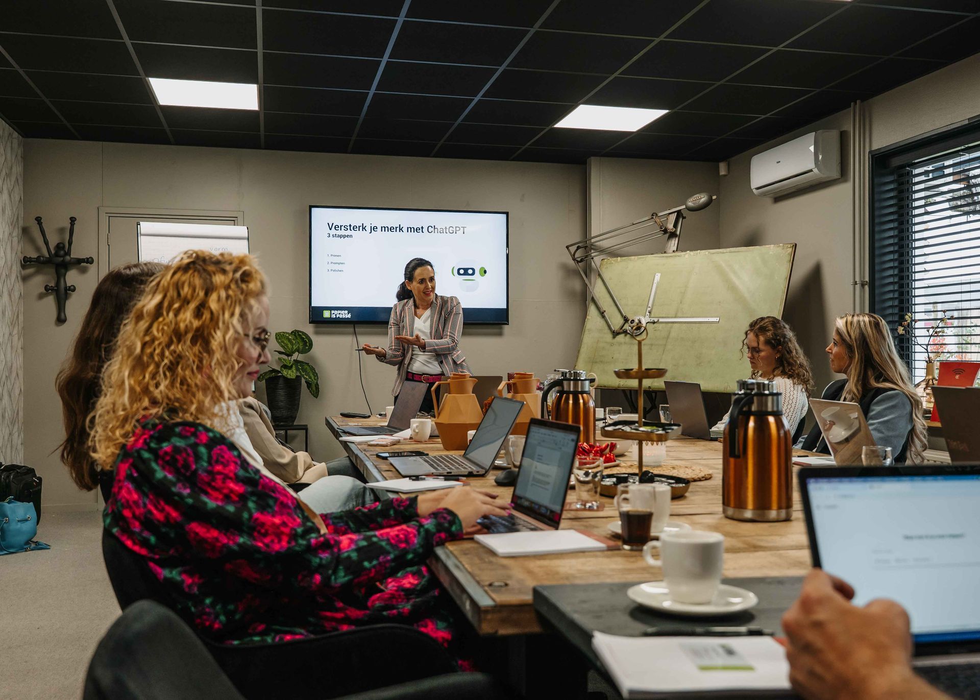 Een presentator staat voor een scherm in een vergaderruimte, terwijl verschillende mensen met laptops aan een houten tafel zitten.