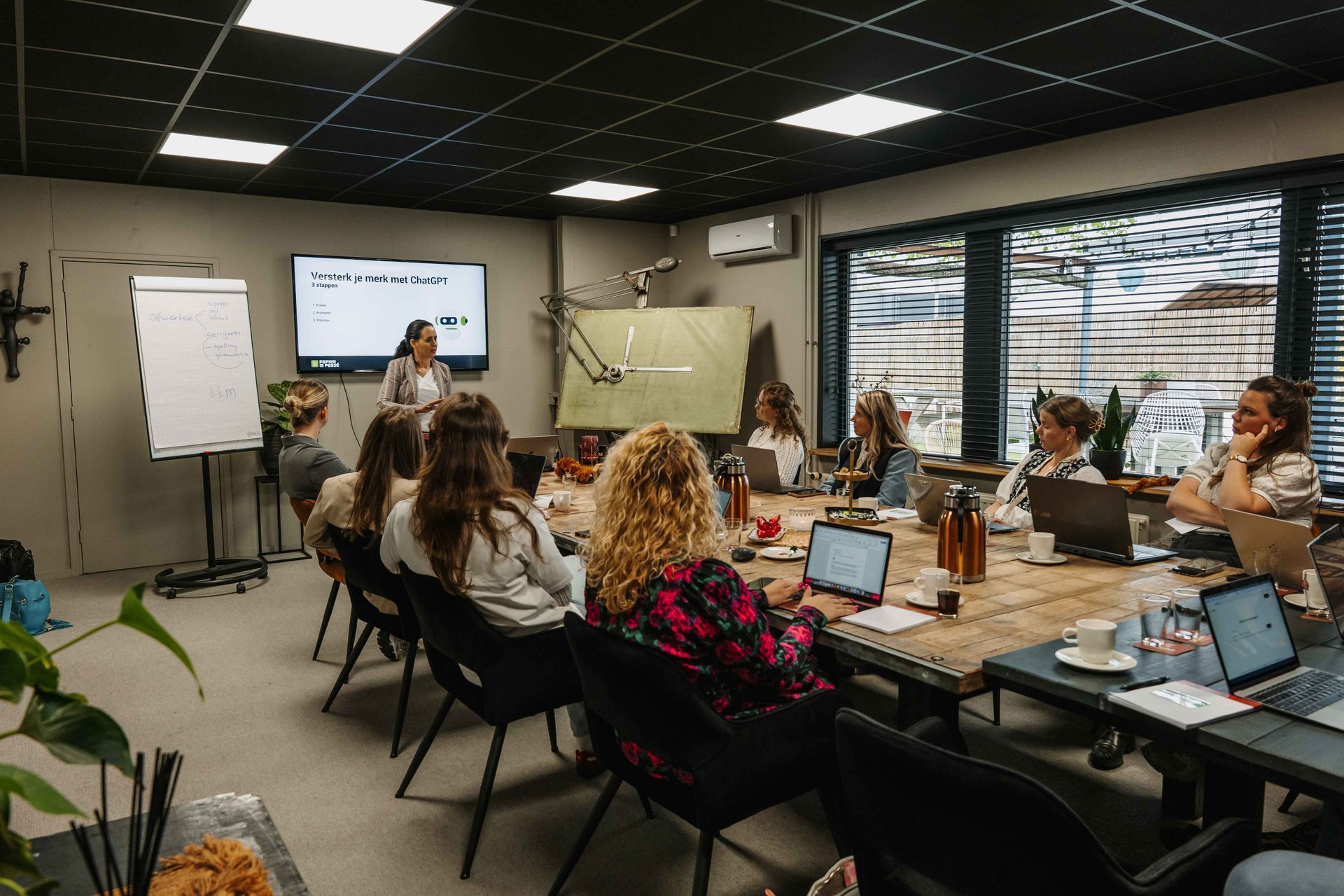 Een groep mensen zit rond een grote houten vergadertafel in een modern kantoor en luistert naar een presentatie.