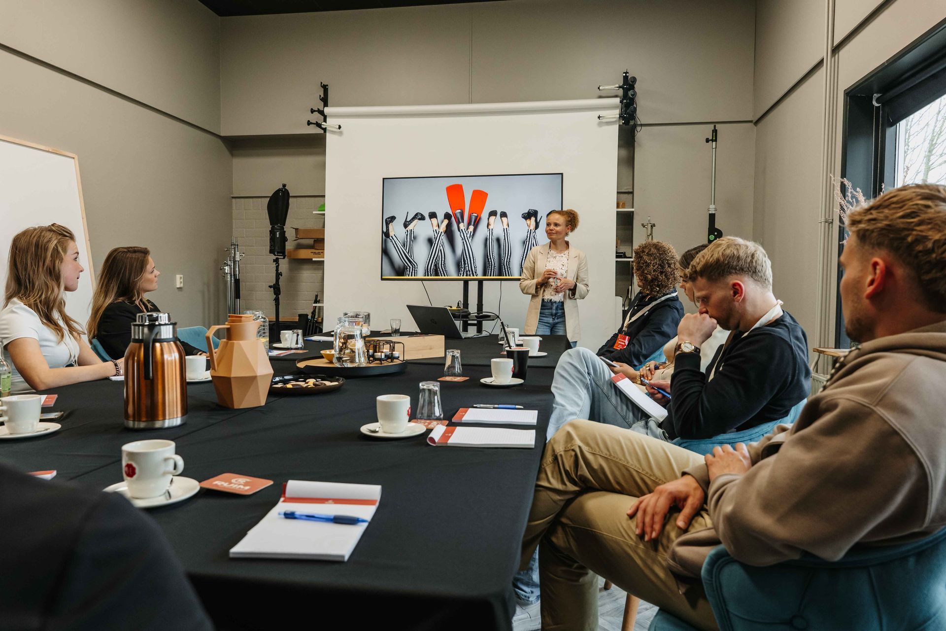 Een groep mensen zit rond een grote tafel in een lichte studio en bekijkt een presentatie op een scherm.