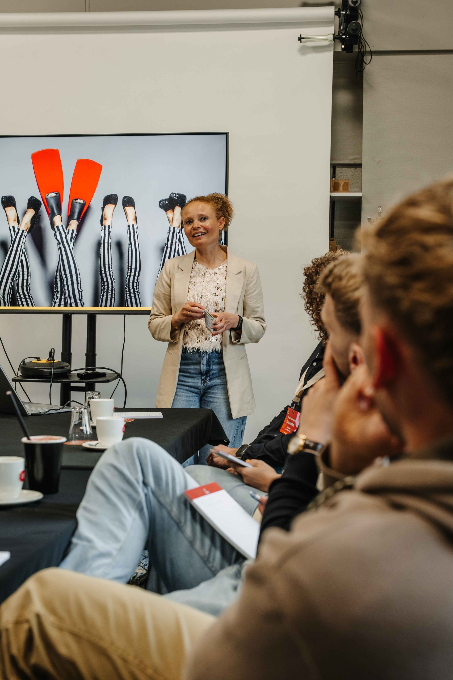 Een presentator spreekt een publiek toe in een studio, met op de achtergrond een scherm waarop modefoto's worden getoond.