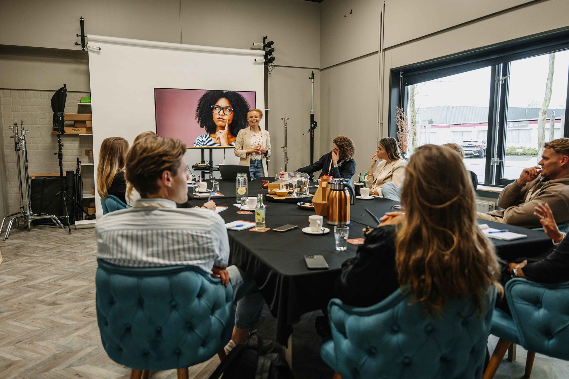Een groep mensen zit rond een tafel in een lichte studio en bekijkt een digitale presentatie op een groot wit achterdoek.