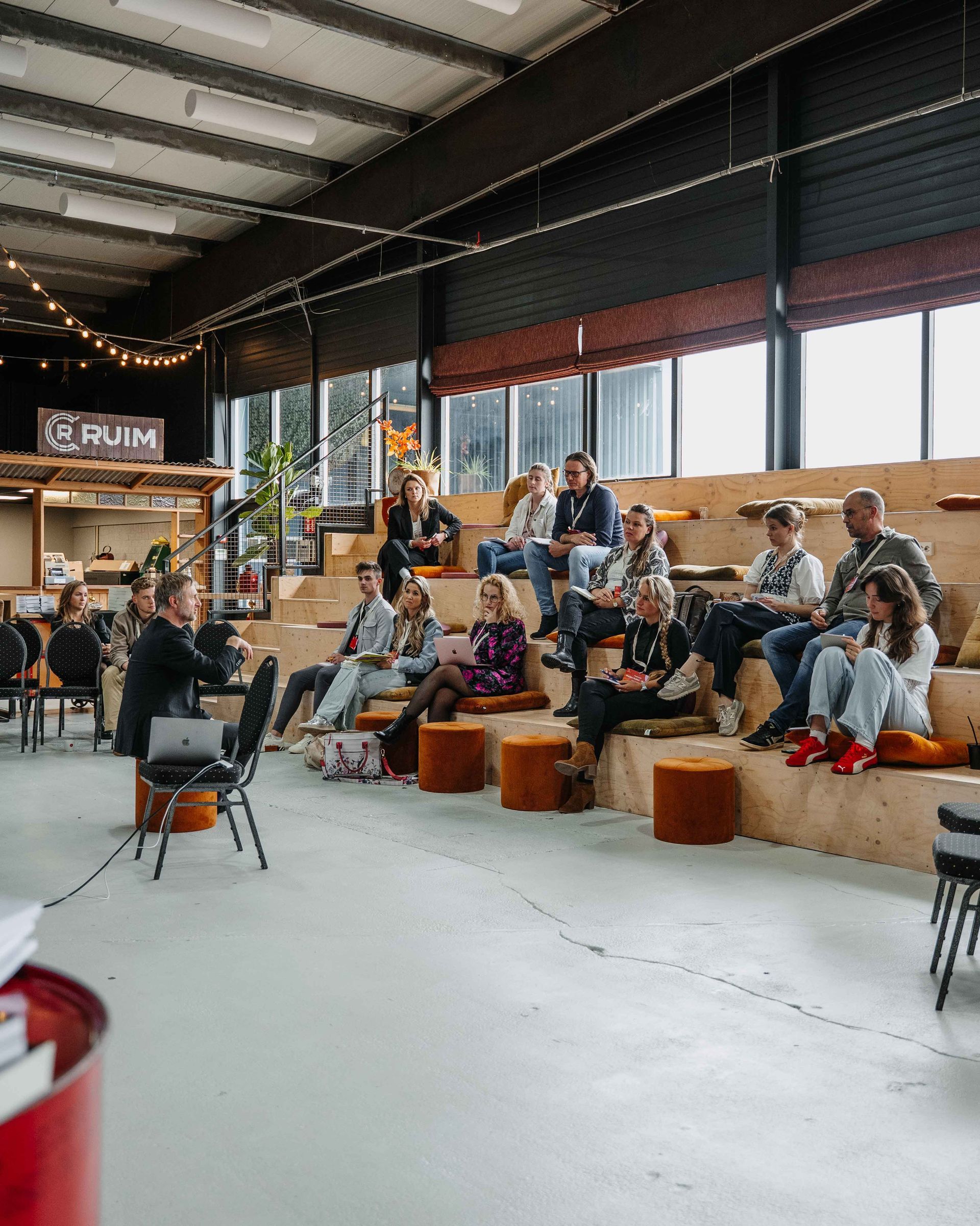 Een spreker spreekt een publiek toe dat op houten trappen zit in een ruime, moderne zaal in industriële stijl.