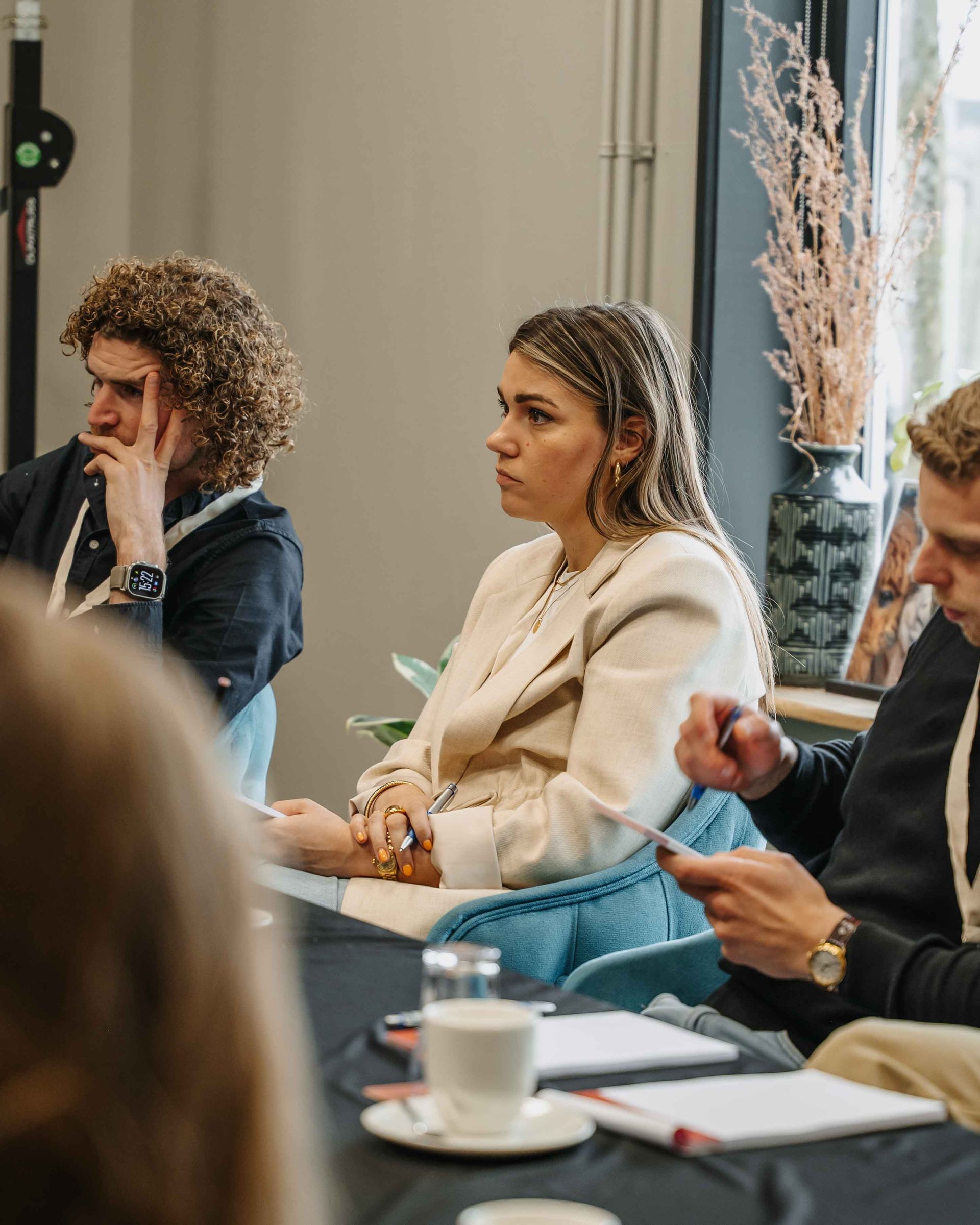 Drie mensen zitten tijdens een vergadering aan een tafel, aandachtig luisterend en aantekeningen makend in een professionele binnenruimte.