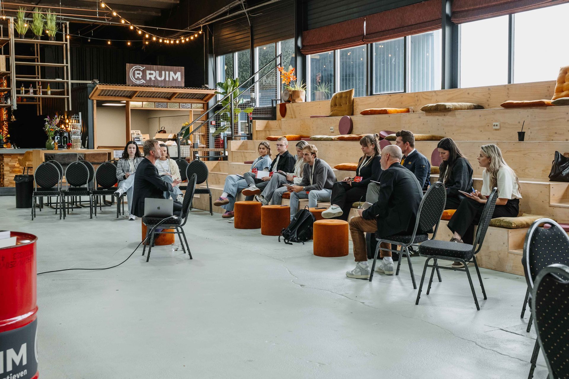 Een spreker geeft een presentatie tijdens een klein seminar binnen in een pakhuis, voor een publiek dat zit op houten tribunes en stoelen.