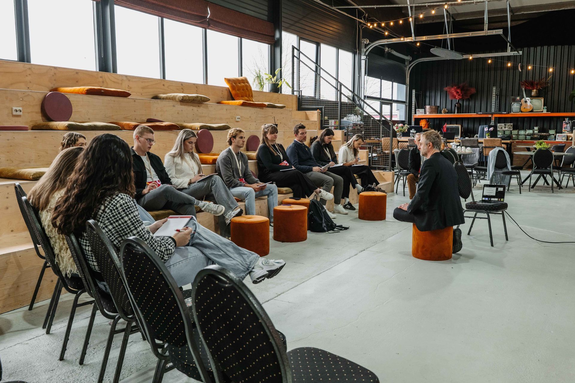 Een groep mensen zit op stoelen en houten zitbanken in een moderne, industriële ruimte en luistert naar een spreker.