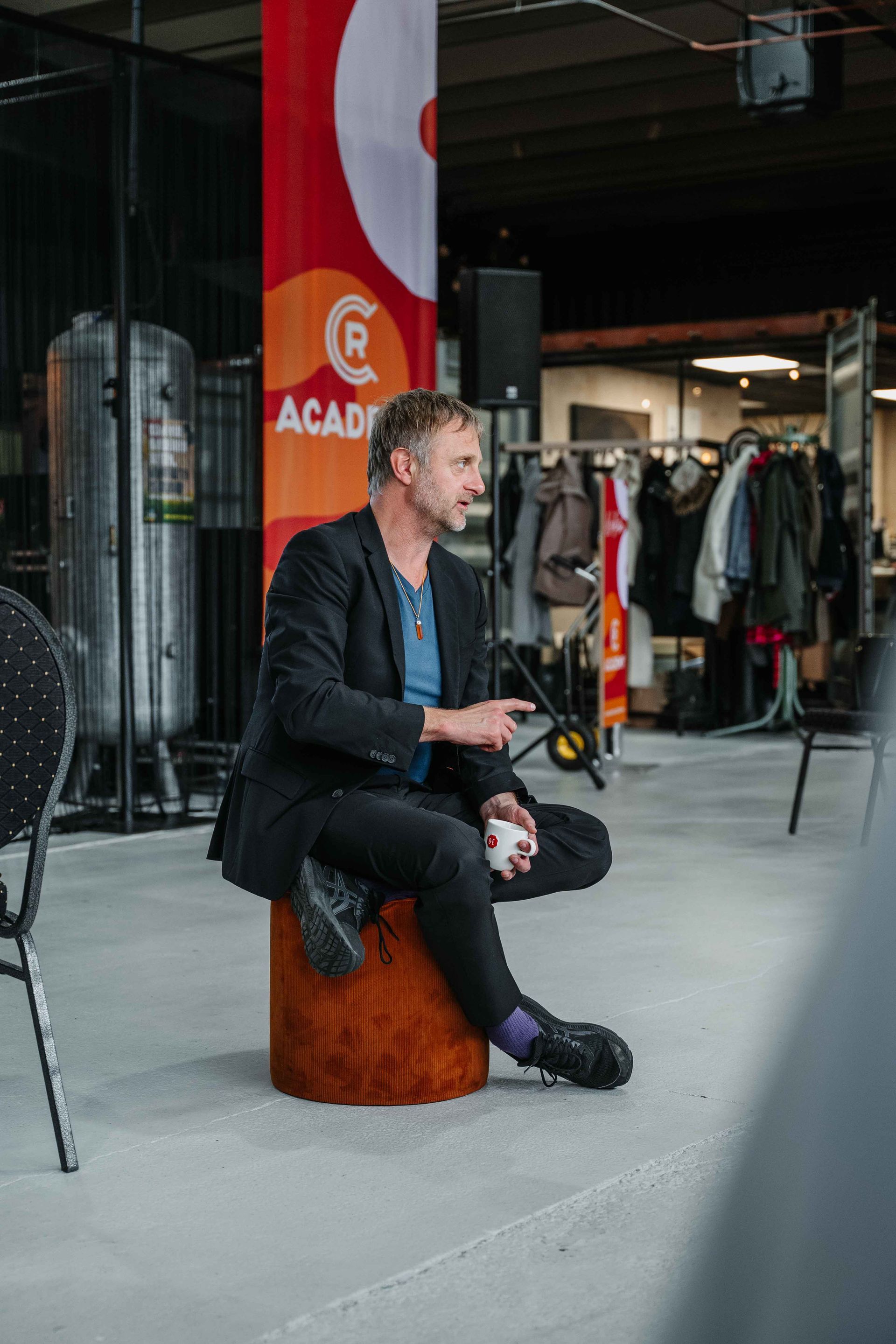 Een persoon in een blazer zit op een houten kruk en gebaart terwijl hij spreekt in een industriële binnenruimte met een rode banner.
