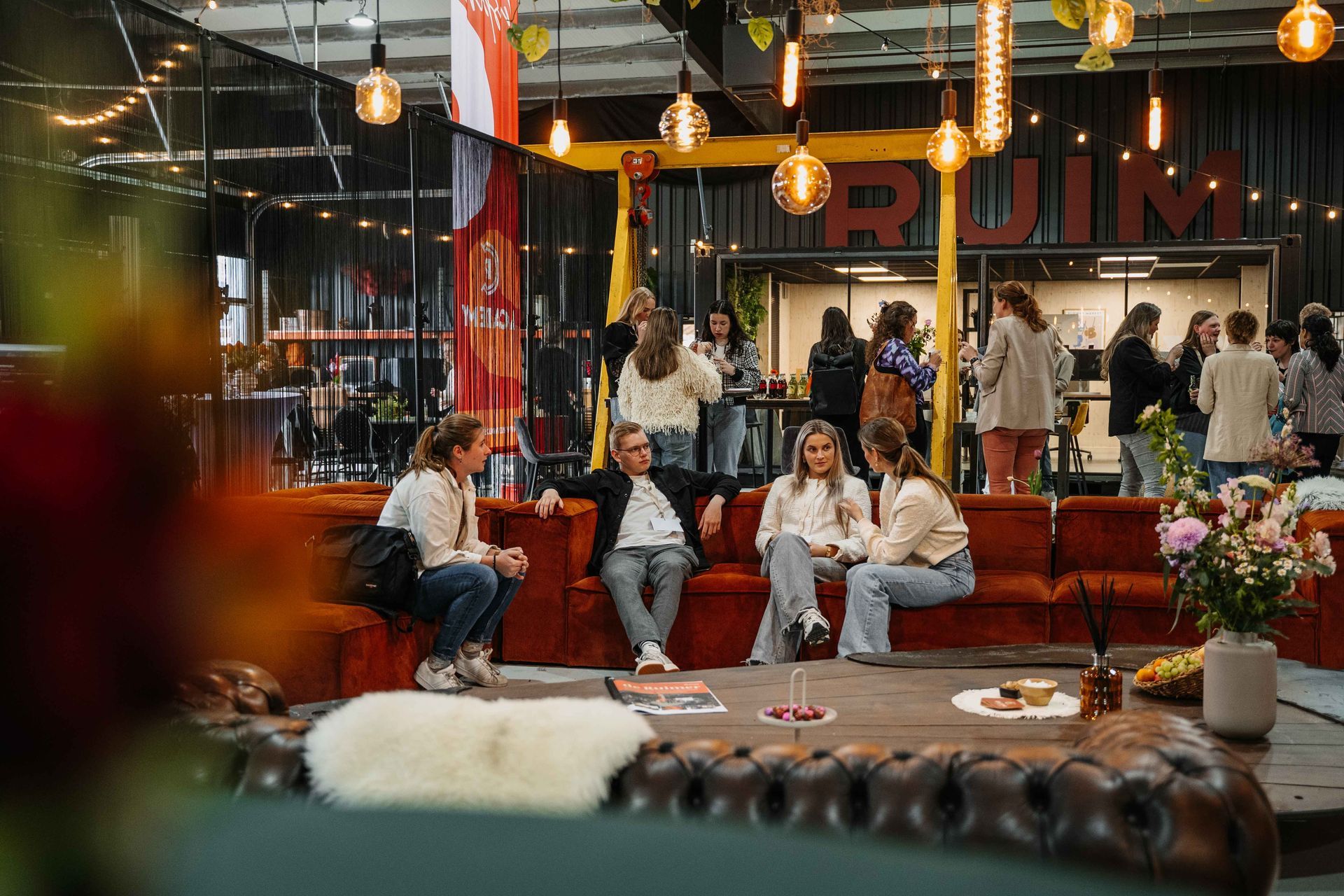 Mensen praten gezellig bij op een roestkleurige bank in een moderne lounge in industriële stijl met warme verlichting en een lage houten tafel.