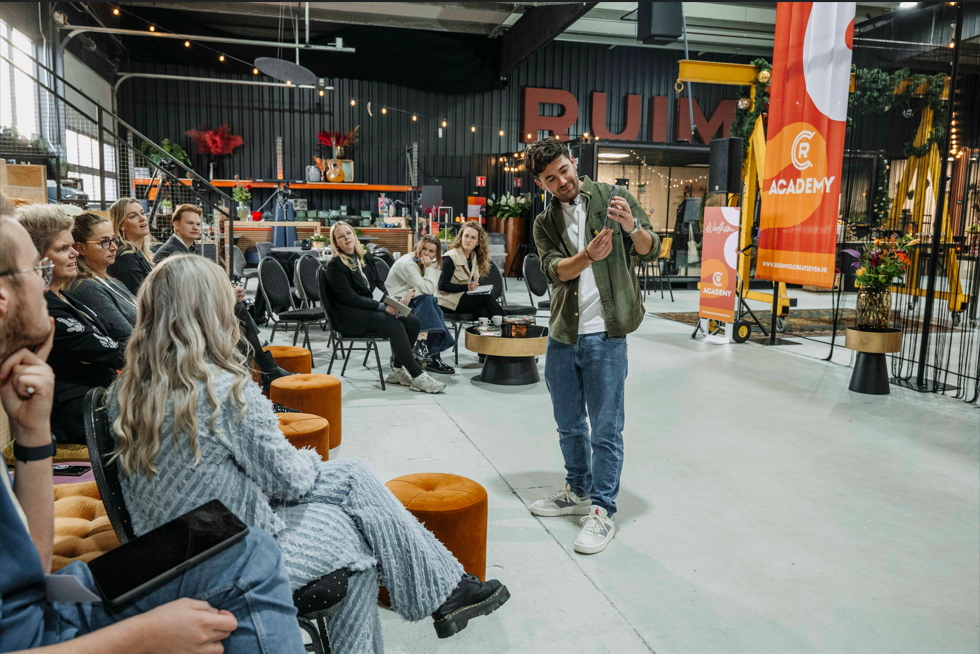 Een man staat en spreekt tot een groep zittende mensen in een studio in magazijnstijl, waaraan een oranje Academy-banner hangt.