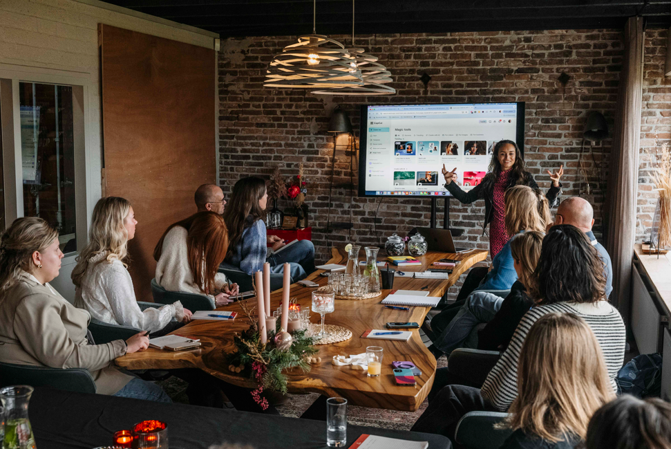 Een vrouw geeft een presentatie aan een groep die rond een grote houten tafel zit in een kamer met een bakstenen muur en een digitaal scherm.