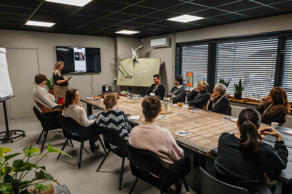 Een groep mensen zit rond een lange houten tafel in een vergaderruimte, terwijl één persoon een presentatie geeft op een scherm.