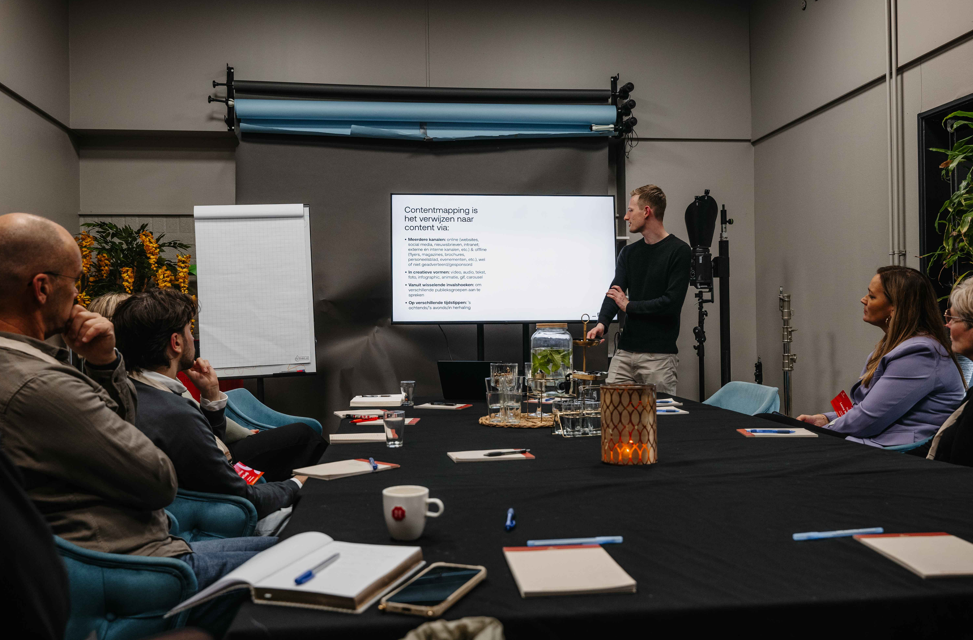 Iemand presenteert een dia aan een groep die rond een vergadertafel zit in een schemerig verlichte, professionele omgeving.