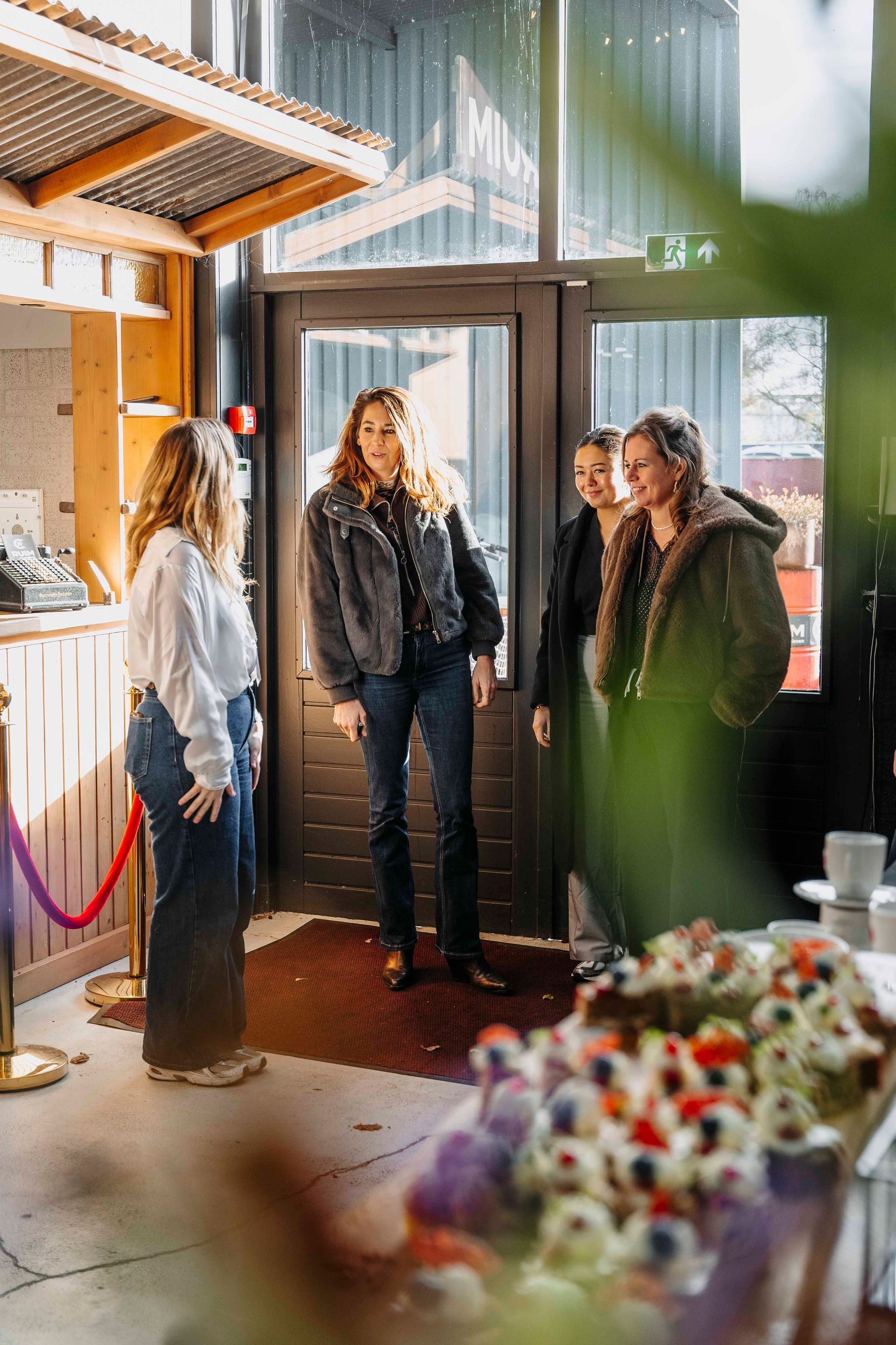 Vier mensen staan ​​bij een deuropening in een schemerig verlichte binnenruimte en praten in de buurt van een houten constructie en een wazige toonbank.