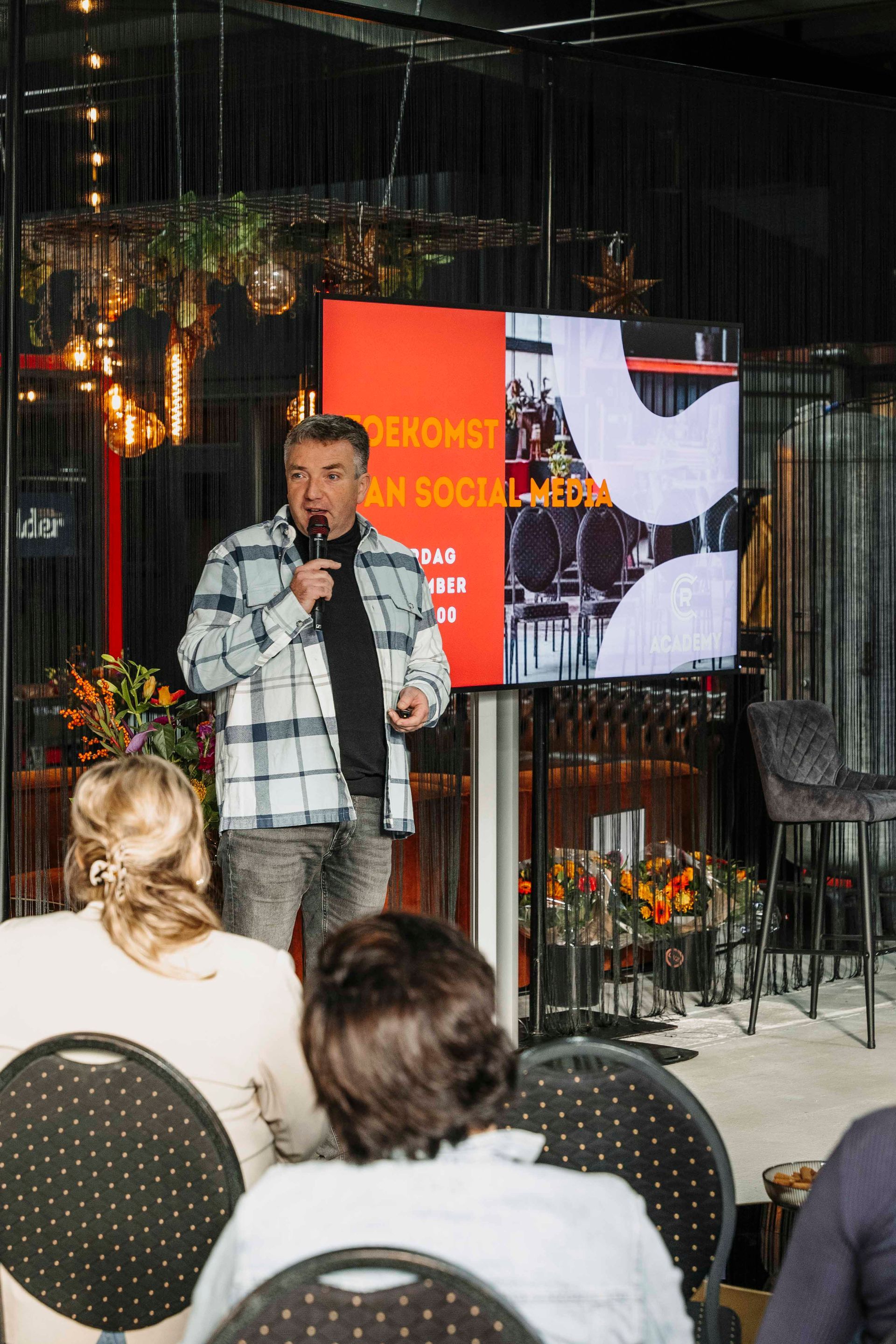 Een spreker in een geruit overhemd geeft een presentatie aan een publiek, met op een digitaal scherm achter hem of haar 