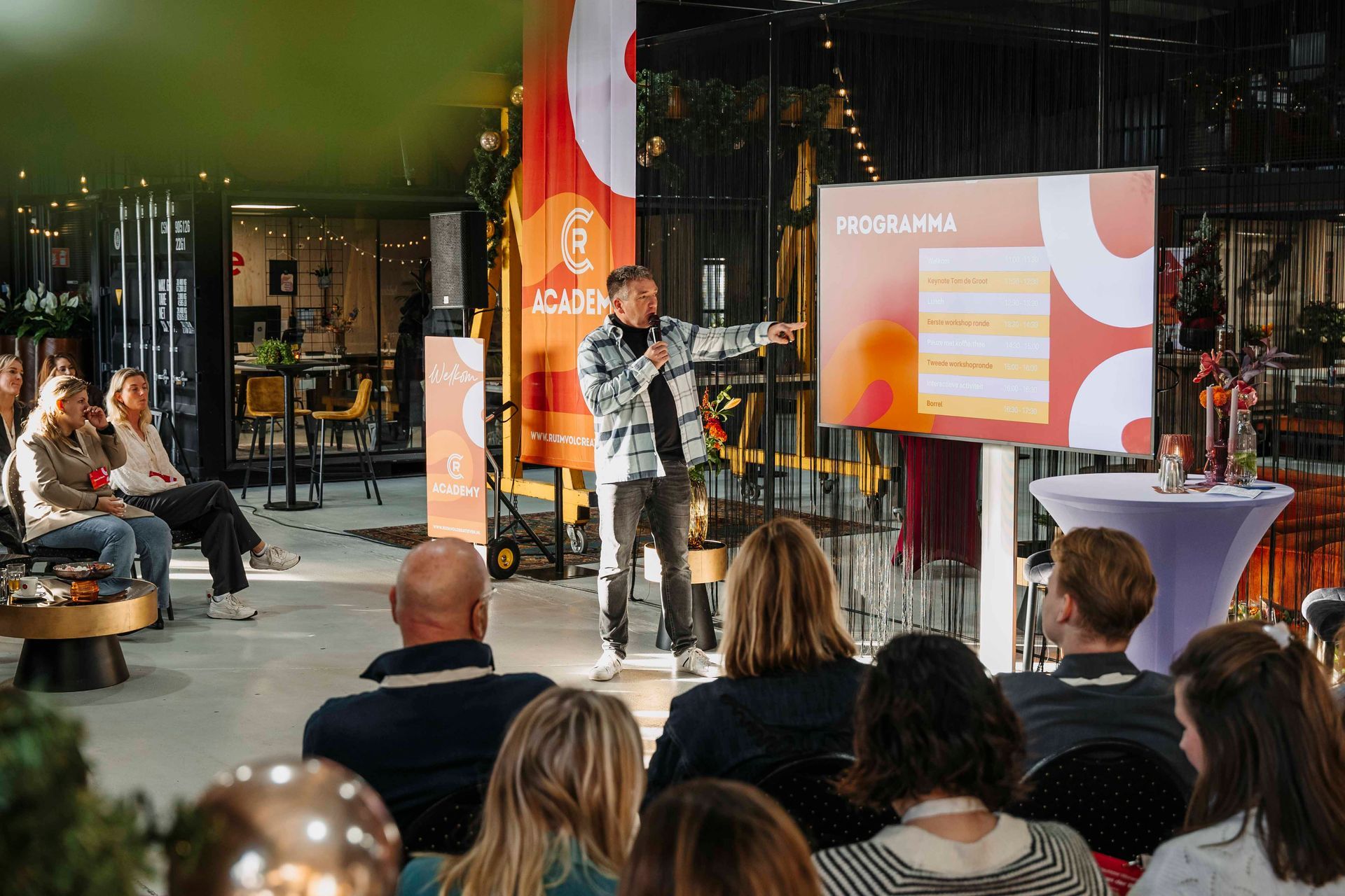 Iemand geeft een presentatie met dia's aan een publiek in een lichte evenementenruimte met warme, oranje tinten in de huisstijl.