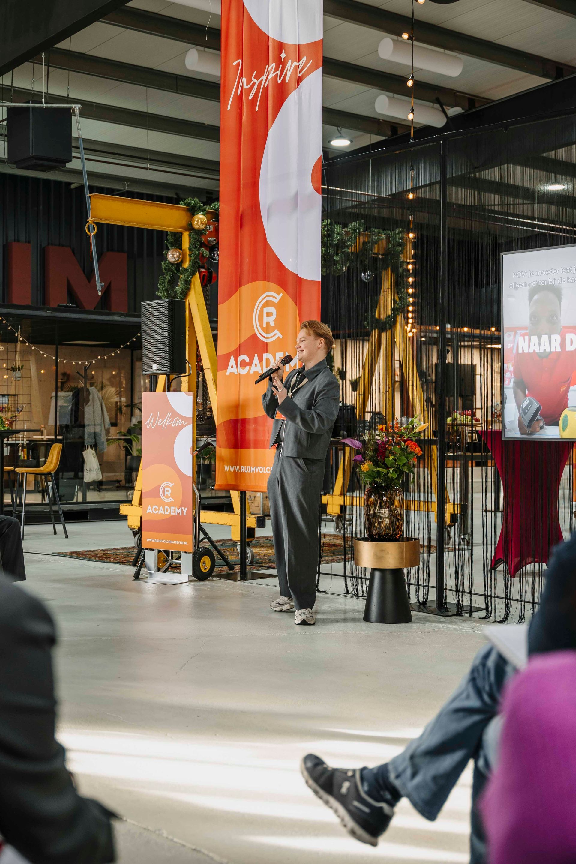 Een spreker spreekt een publiek toe vanaf een podium met een hoog oranje spandoek in een open, moderne binnenruimte.