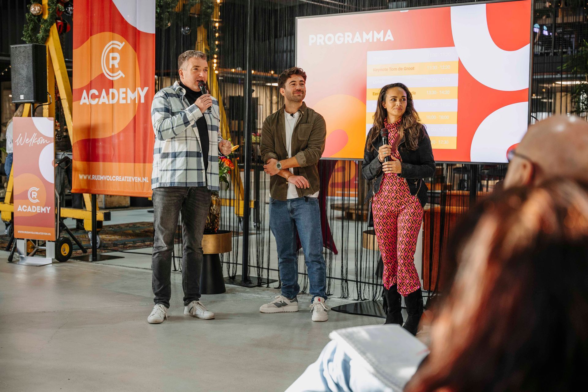 Een spreker staat op een podium met twee personen naast zich in een felverlichte industriële ruimte met evenementbanners met logo.