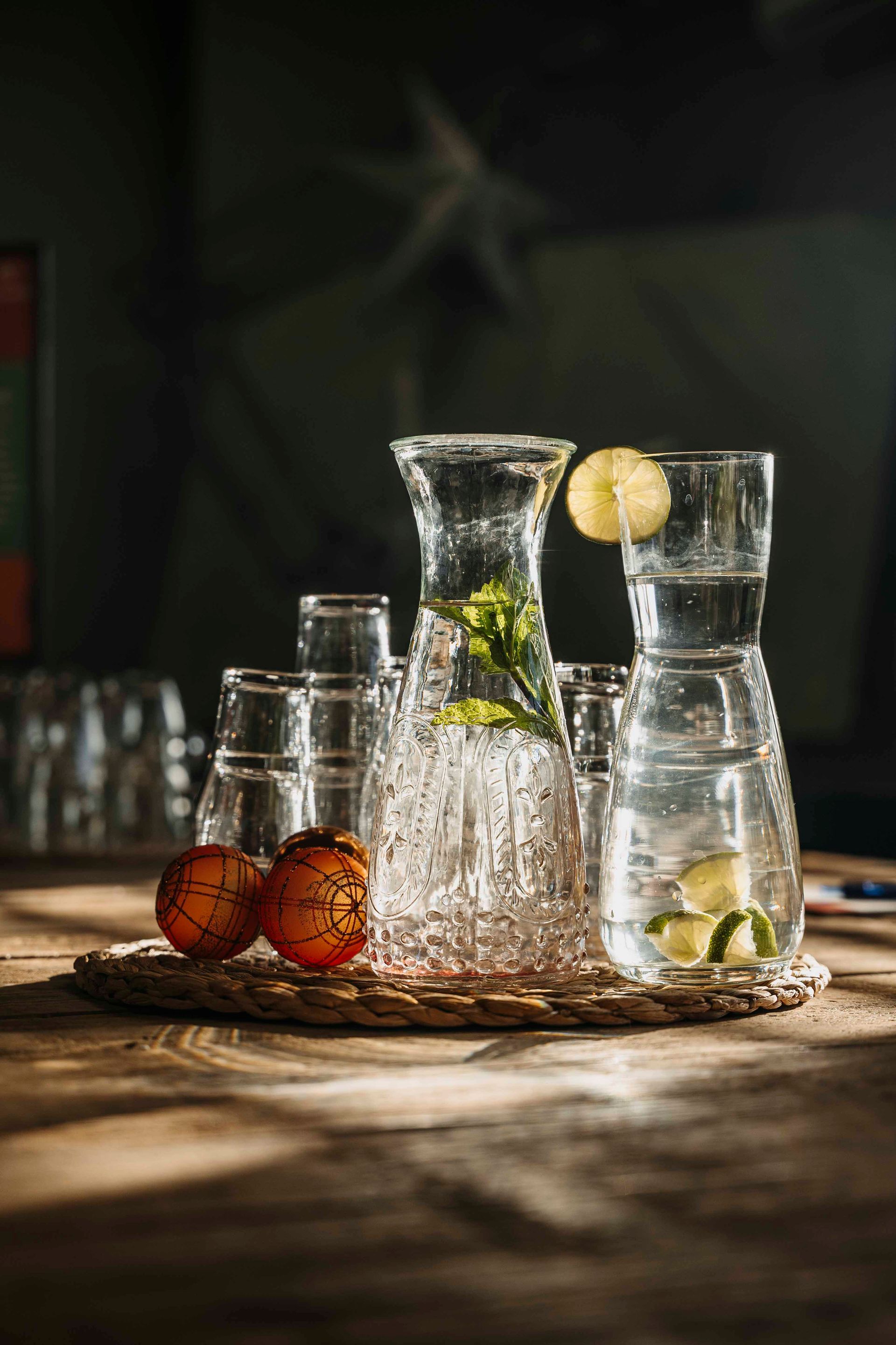 Een karaf van getextureerd glas met een garnering en een glas water met citrussmaak staan ​​op een rieten matje op een houten tafel.