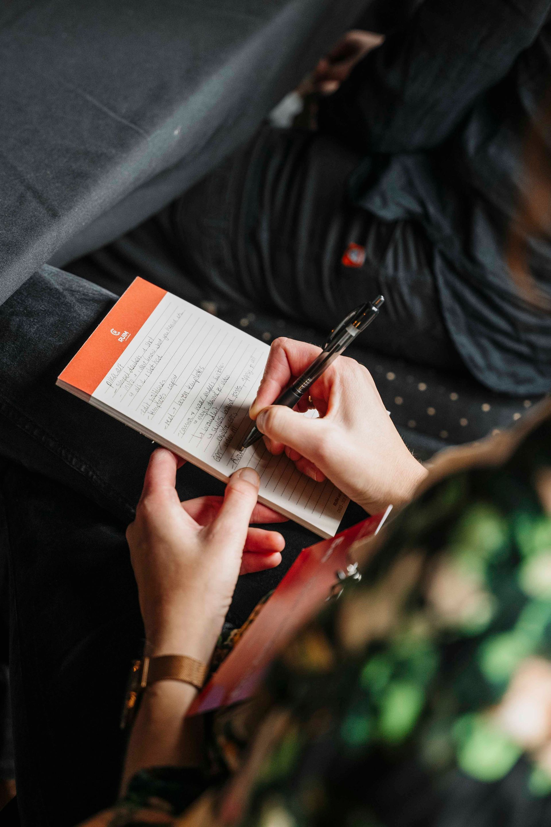 Een persoon schrijft met een zwarte pen in een klein notitieblokje met een oranje bovenkant, dat hij of zij in de hand houdt.