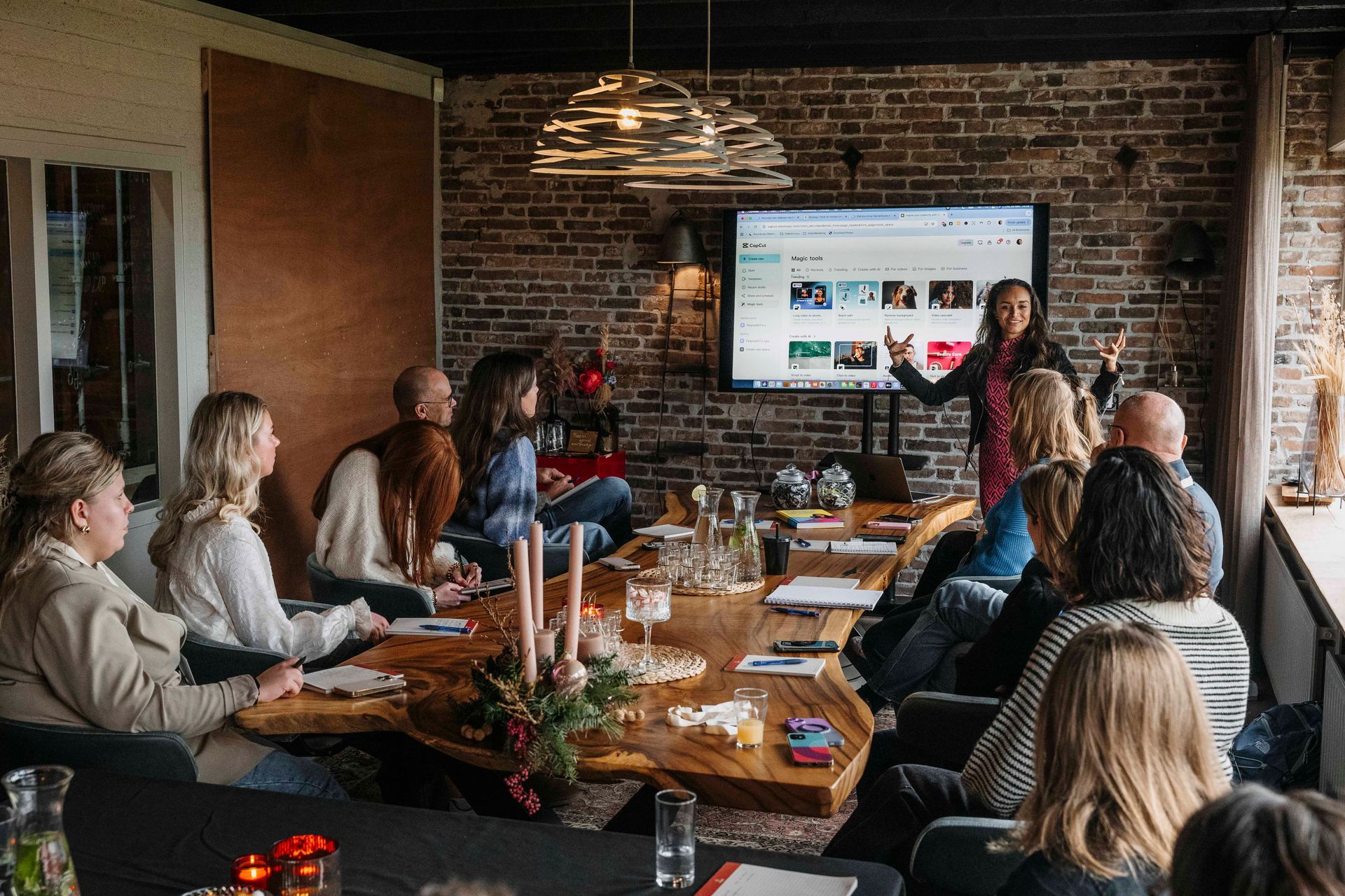 Een groep mensen zit rond een houten tafel in een kamer met bakstenen muren en kijkt naar een presentatie op een groot scherm.